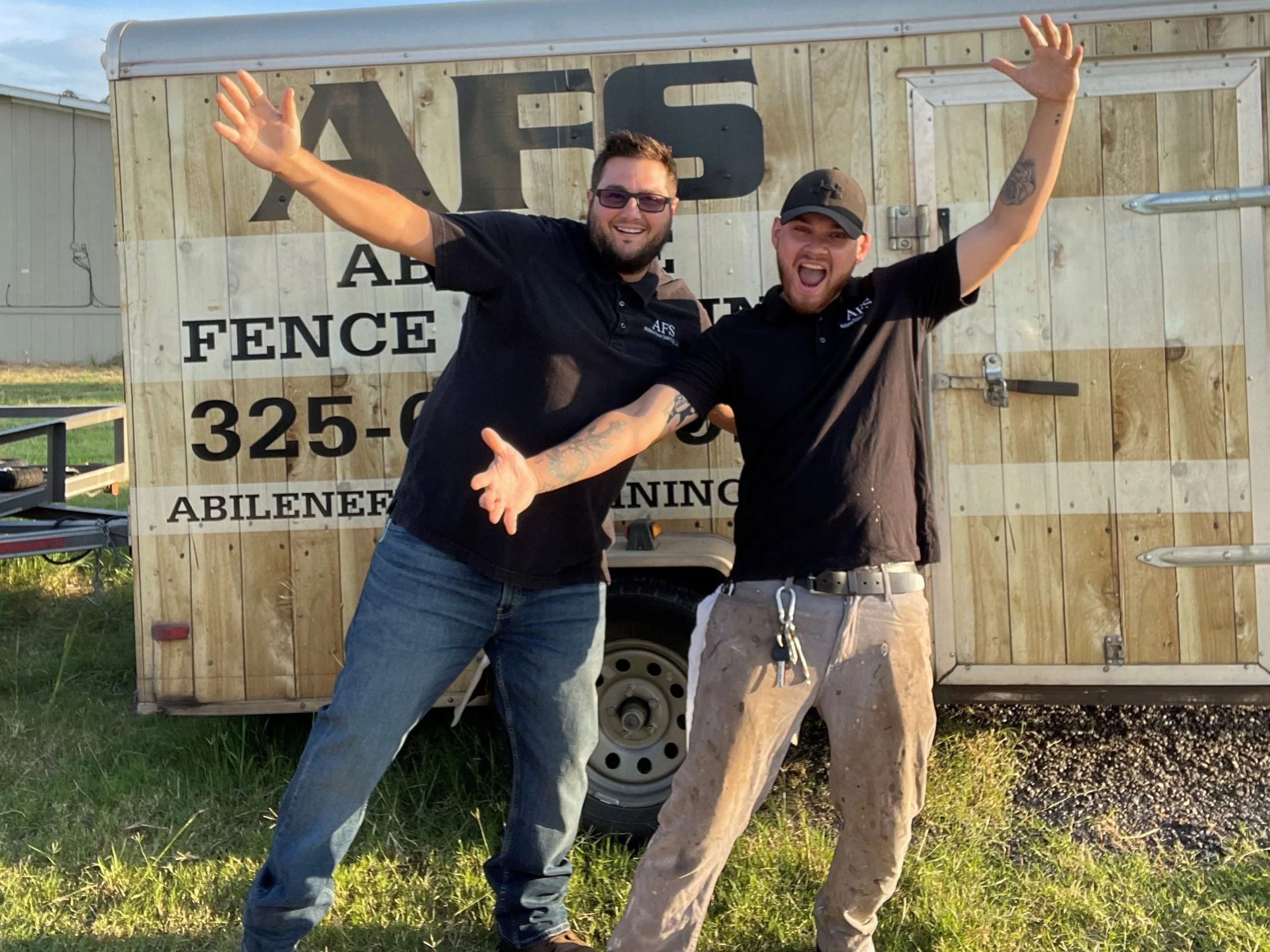 Two men smiling and waving in front of a wooden fence trailer. One man has a beard, glasses, and is wearing a black shirt. The other man has a beard, a cap, and a black shirt. The trailer has the words 'ABLANE FENCE' and a phone number printed on it.