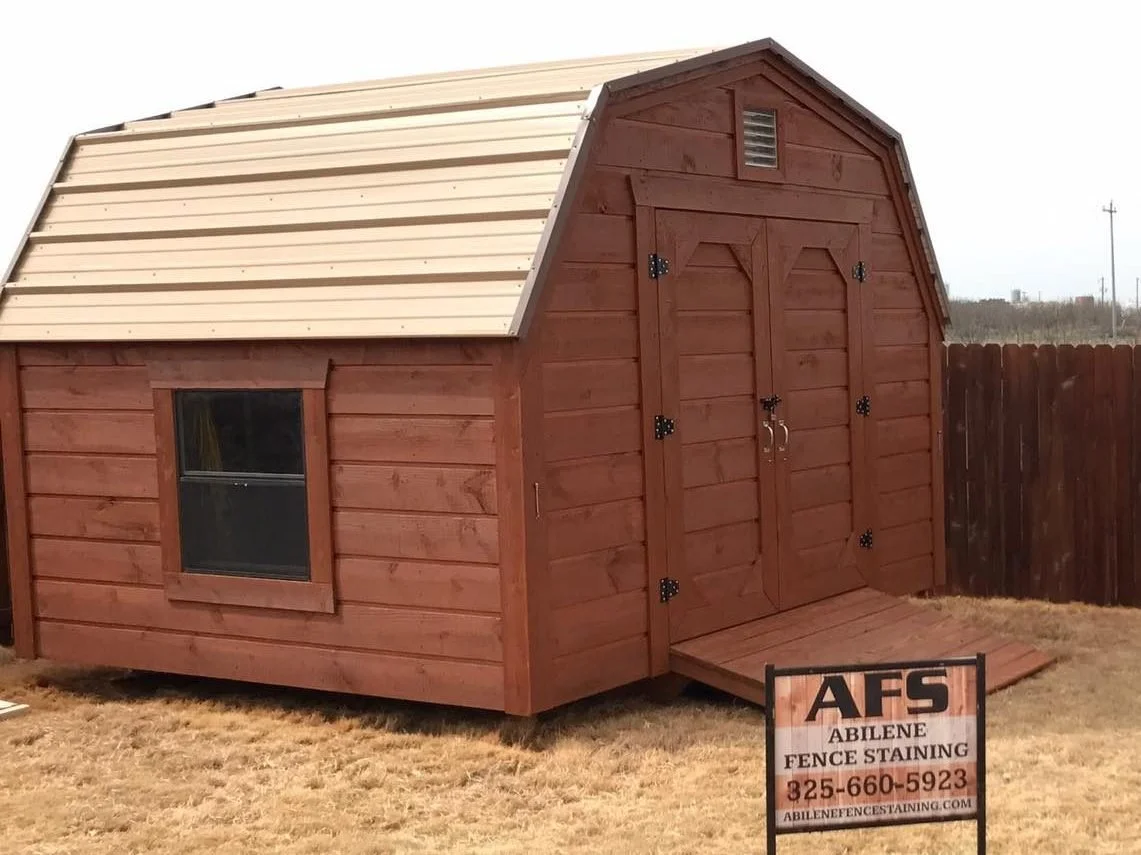 A small wooden shed painted in reddish-brown color with a double door and a small window on the side, situated on a patch of grass with a wooden fence in the background. There is a sign in front of the shed reading 'AFS Abilene Fence Staining' with a