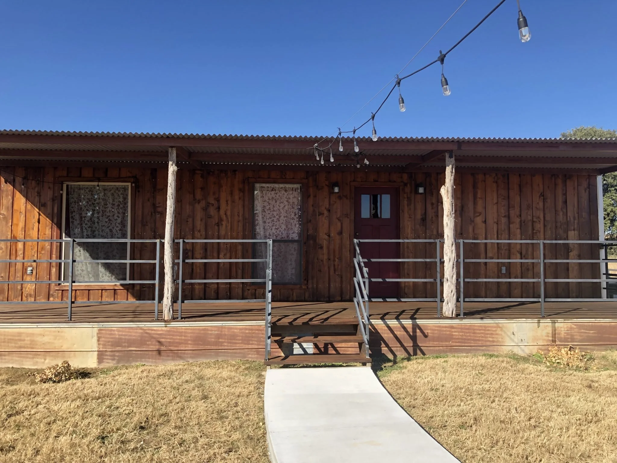 A small rustic wooden house with a corrugated metal roof, a red door, two windows with floral curtains, a small porch with steps, and a string of outdoor string lights overhead, with grassy yard and clear blue sky.