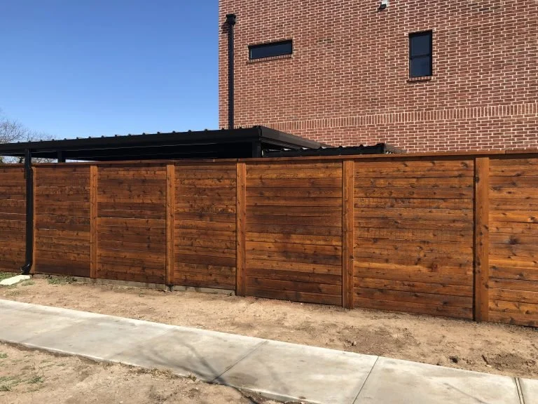 Wooden privacy fence in front of a brick building with small rectangular windows, sidewalk and clear blue sky.