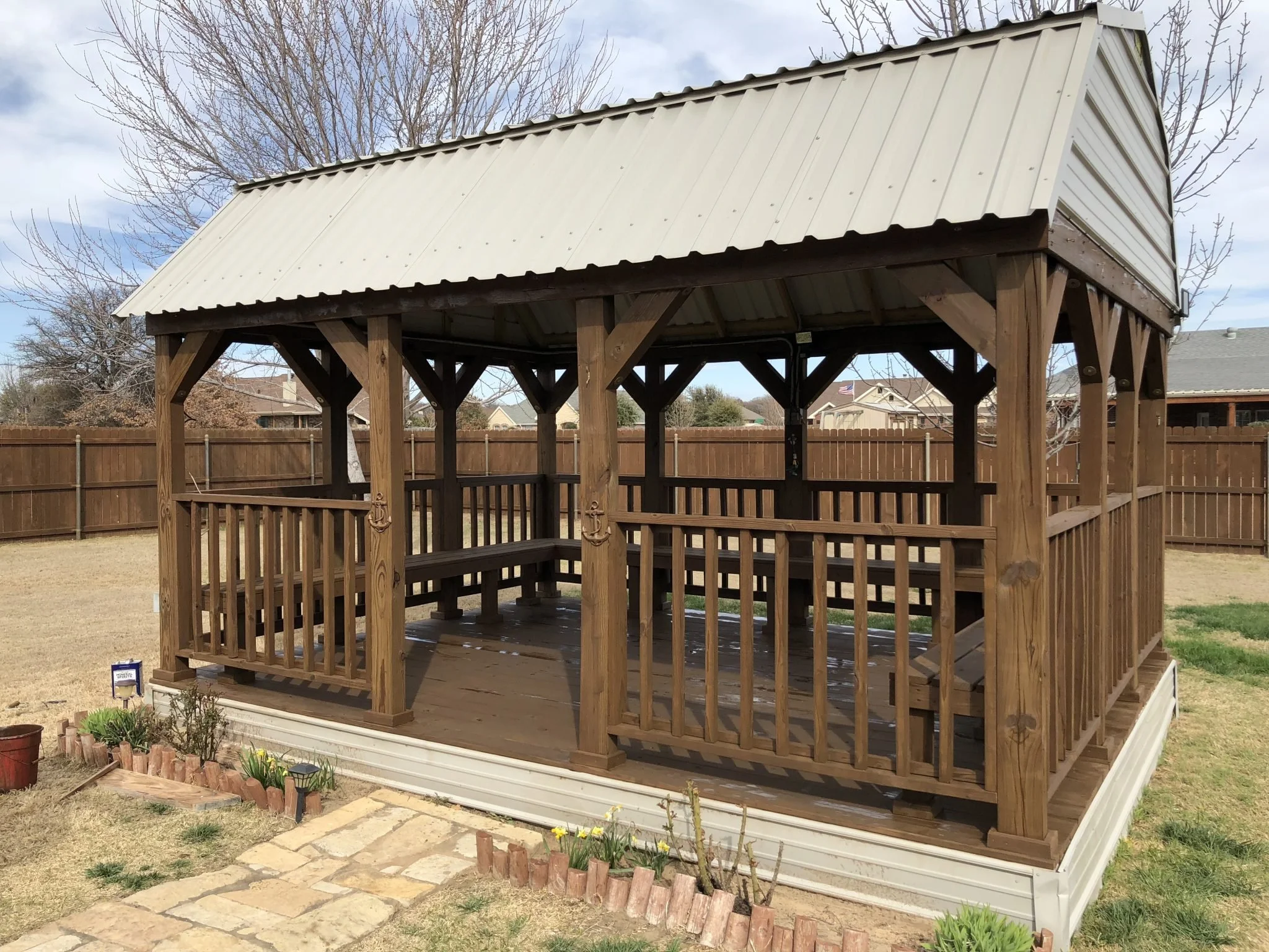 Wooden pergola with a white metal roof, situated in a backyard with a fence and a small garden with flowers and a stone path.