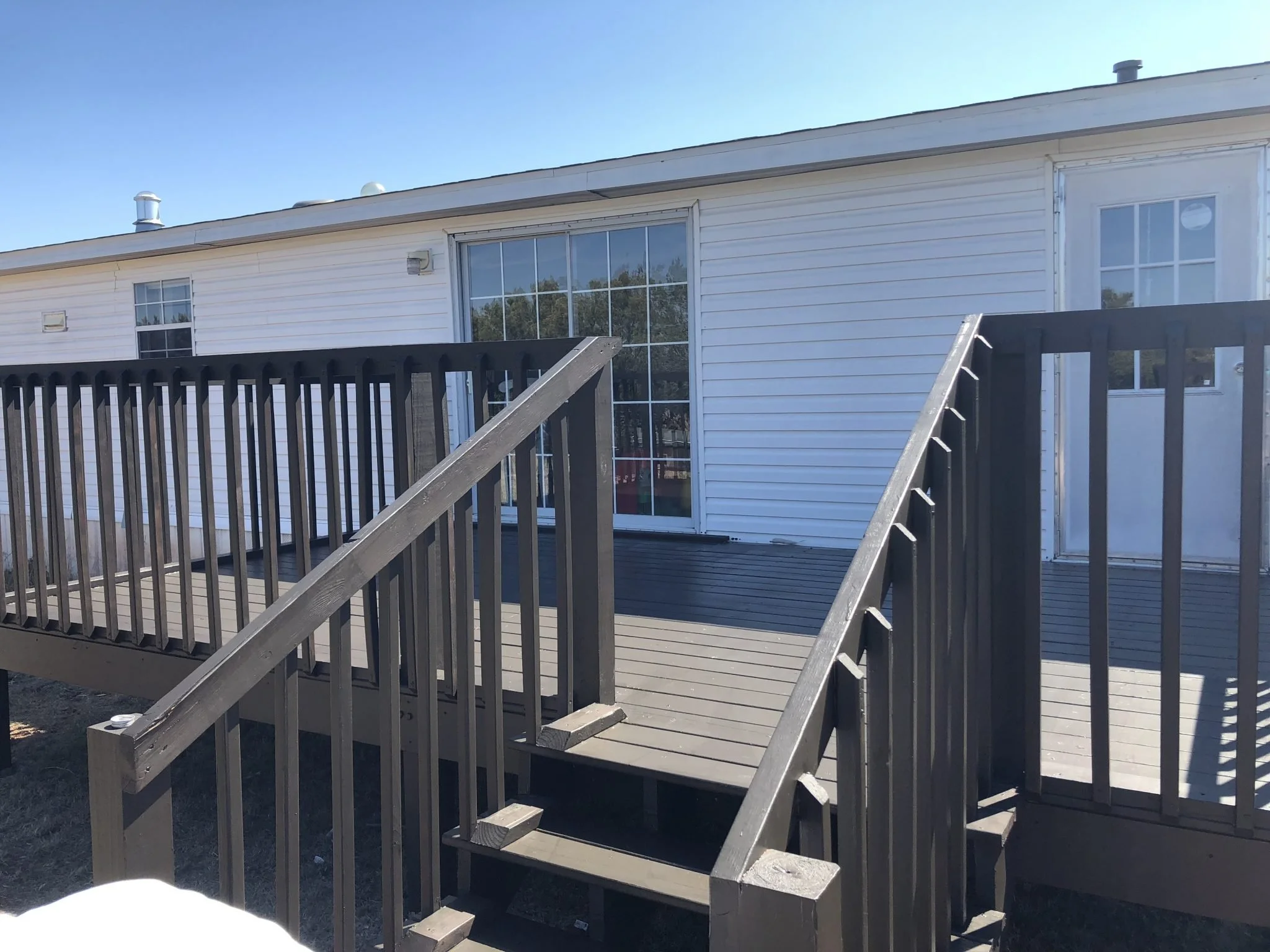 A wooden deck with stairs leading up to a sliding glass door on a white house. The house has a white exterior with horizontal siding, and a window can be seen to the right of the door. The sky is clear and blue.