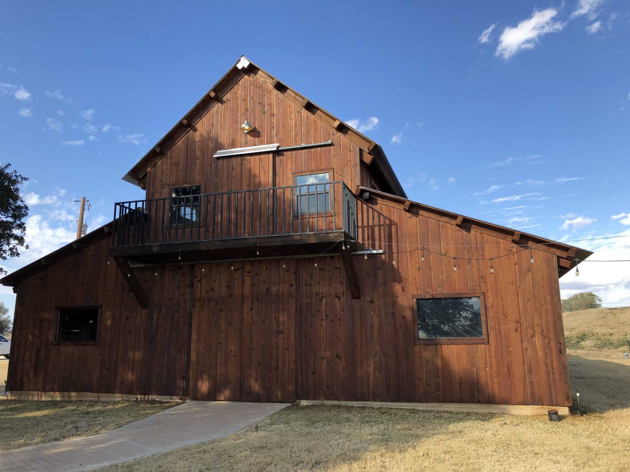 A wooden house with a triangular roof, a balcony with a metal railing, and several windows. The house is set against a blue sky with some clouds and is surrounded by a grassy yard with a paved walkway.