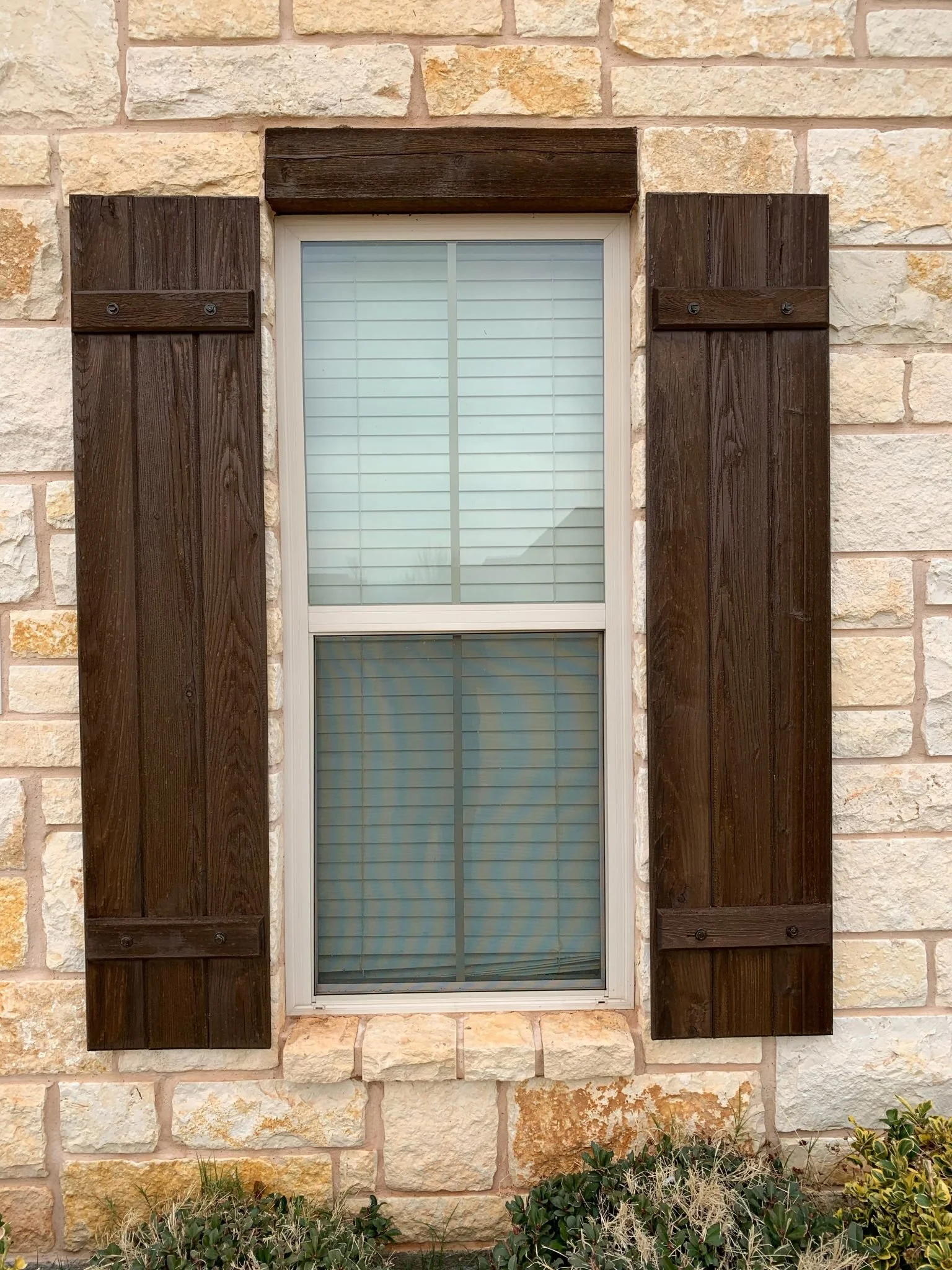 A window with white blinds flanked by dark brown wood shutters on a light beige stone wall with some green plants at the bottom.