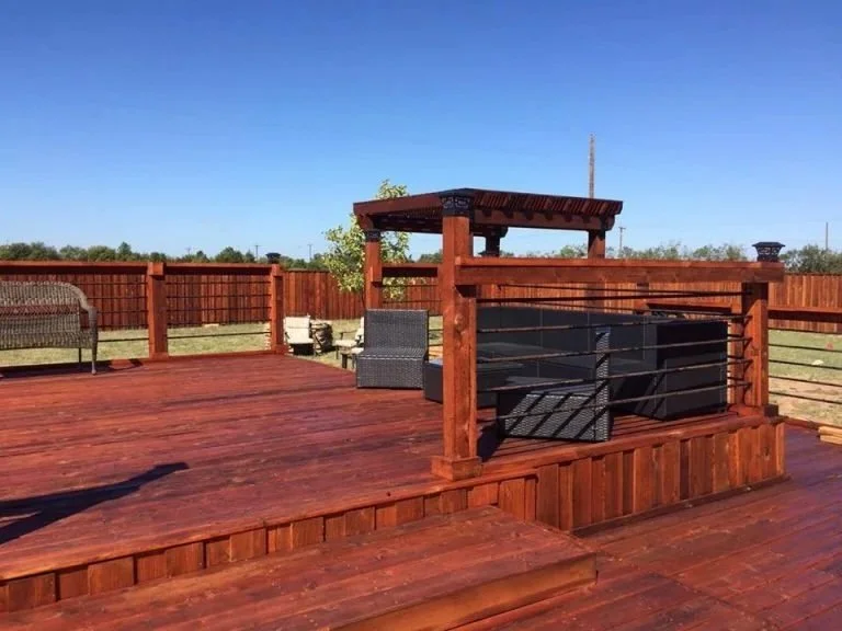 Wooden deck with outdoor furniture and a barbecue grill under a covered pavilion, fenced backyard with open sky.