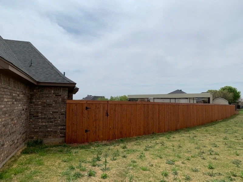 A backyard with a wooden privacy fence stretching along the grass, with a brick house on the left and a cloudy sky overhead.
