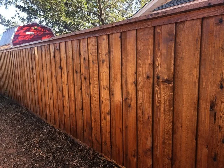 A long wooden privacy fence with vertical planks, located along a backyard with trees in the background and an outdoor umbrella partially visible.