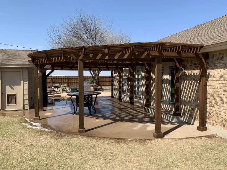 A wooden outdoor pergola with a lattice roof over a concrete patio, attached to a brick house. There is outdoor furniture and a tree in the yard, with a clear blue sky.