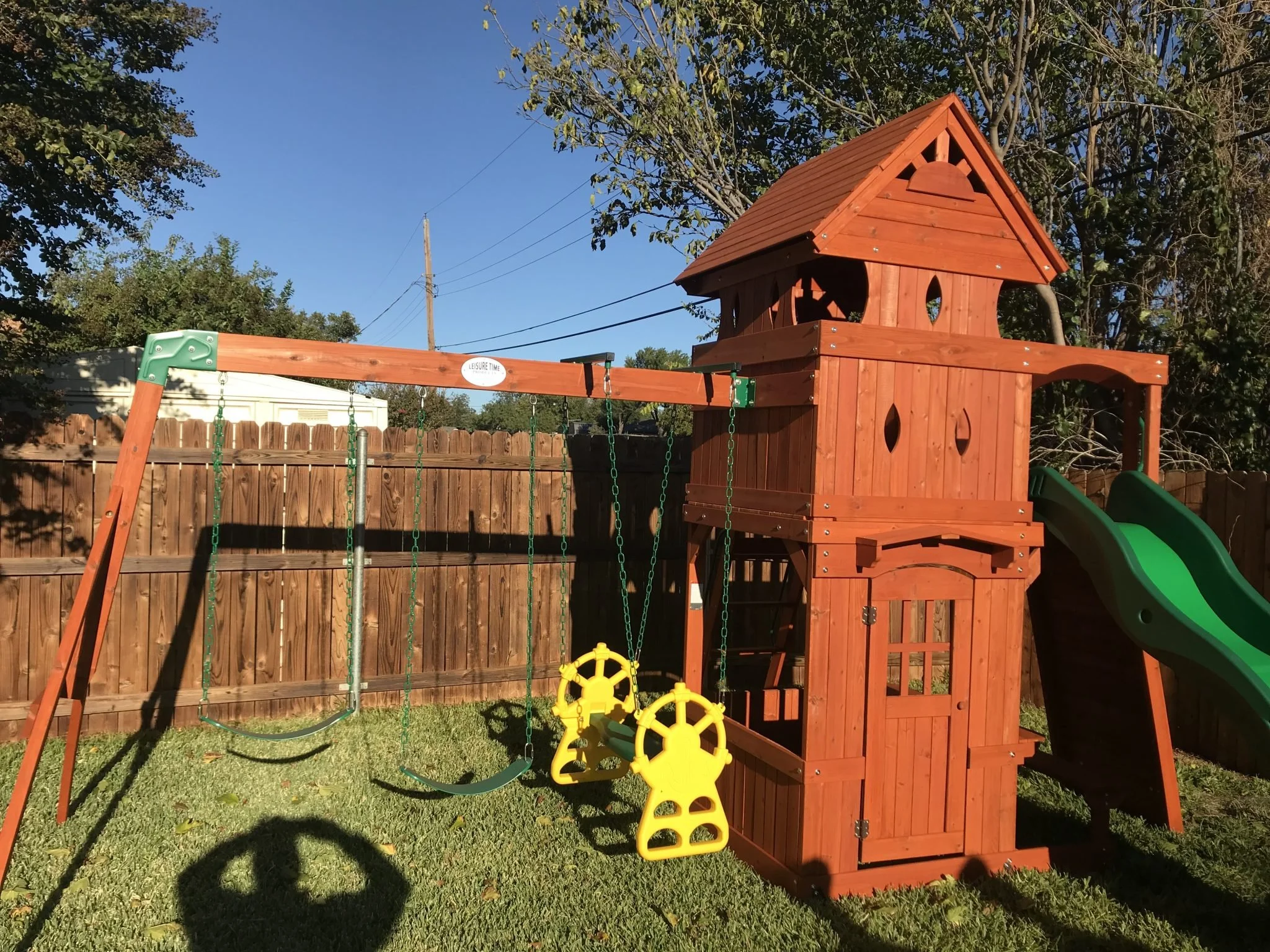 A backyard playground with a wooden playhouse, swings, and a slide, surrounded by a wooden fence and trees, on a sunny day.