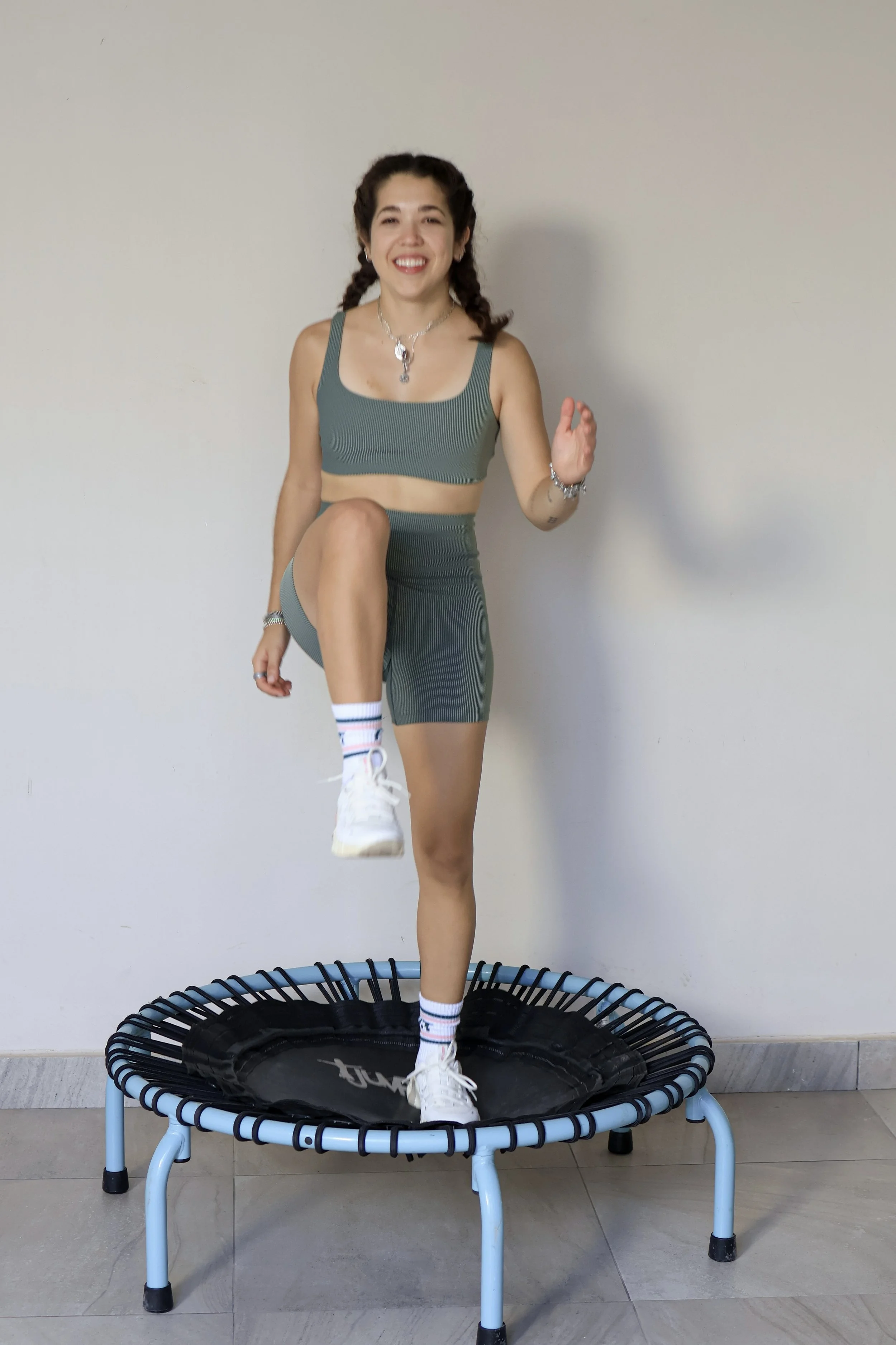 A smiling woman with braided hair is standing on a small trampoline indoors. She is wearing a green sports bra and matching shorts, white sneakers, and striped socks. She has jewelry on her neck and wrists.