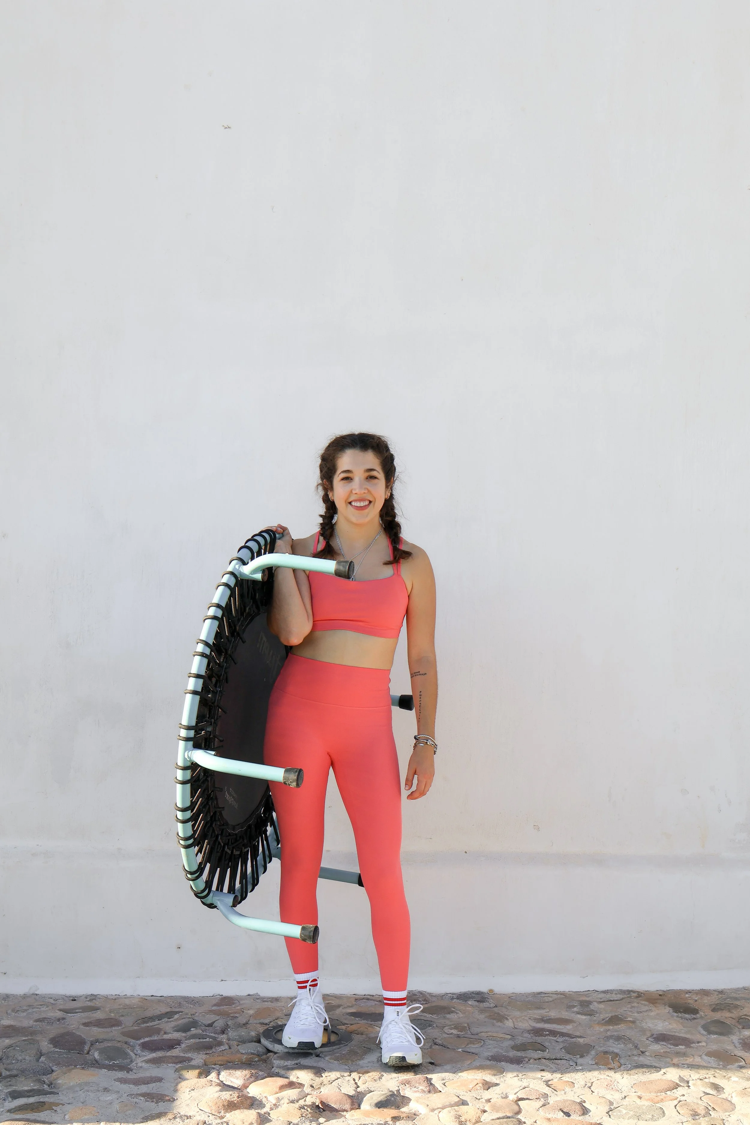 A young woman in pink athletic wear and white sneakers standing against a plain white wall, holding a fitness trampoline with a black net. She has braided hair, a big smile, and is wearing bracelets and a necklace.