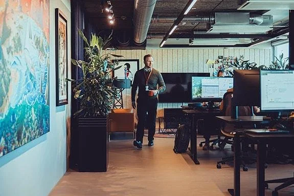 Man walking through a modern open-plan office with desks, computer monitors, plants, and artwork on the walls.