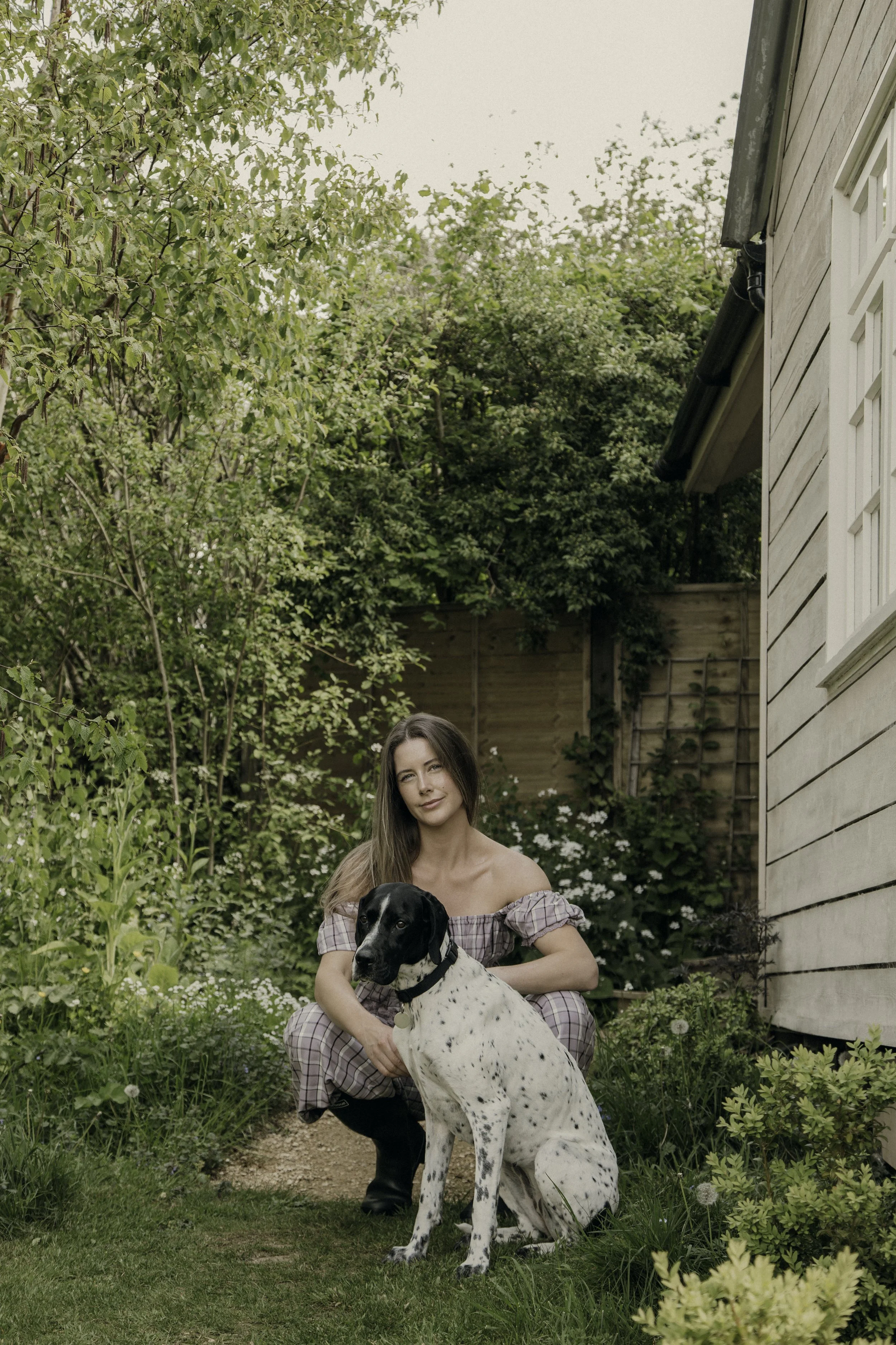 A woman with long brown hair wearing a plaid dress crouches in a garden, holding a large black and white dog. The garden is lush with green plants and white flowers. A wooden fence and a house wall are visible in the background.