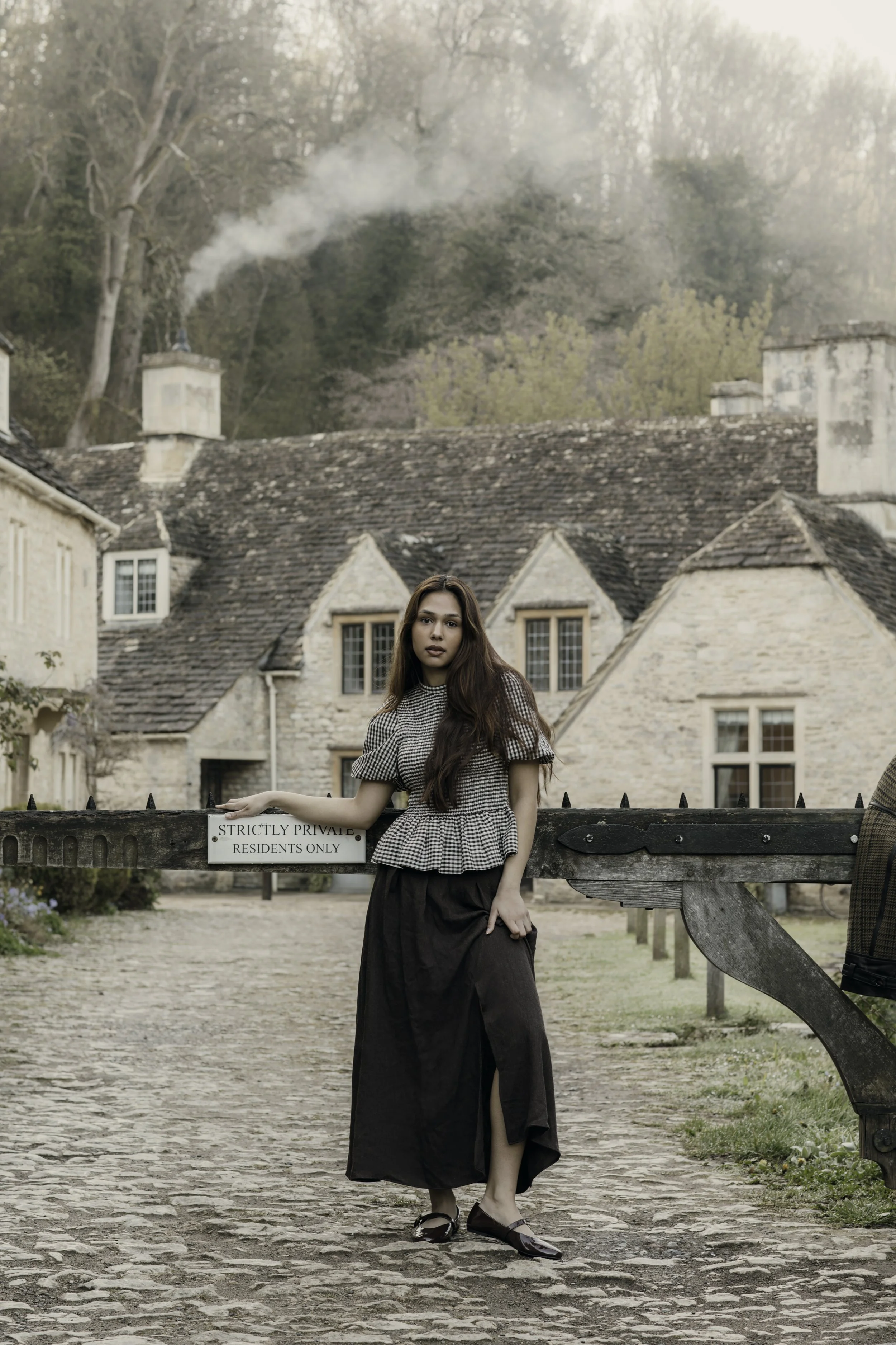 A young woman with long dark hair stands in front of a historic stone building with a cobblestone pathway. She is wearing a black and white patterned top, a long black skirt, and shiny black shoes. She is leaning against a wooden gate with a sign that reads "Strictly Private, Residents Only". In the background, there are trees and a stone house with chimney stacks, and smoke is rising from one of the chimneys.