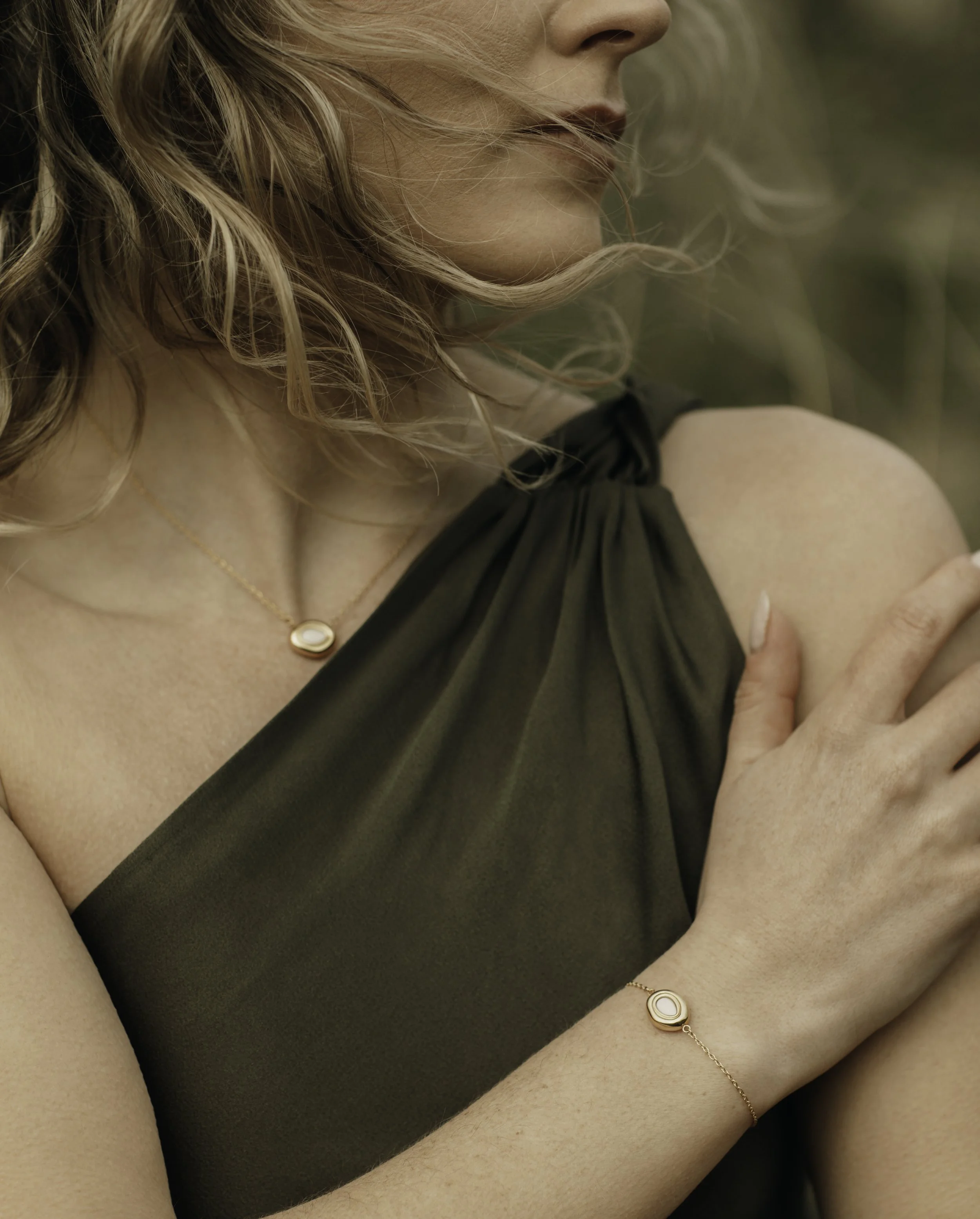 Close-up of a woman's face, shoulder, and arm, with blonde curly hair, wearing a black sleeveless dress, a delicate gold necklace with a circular pendant, and a matching bracelet, outdoors with blurred background.