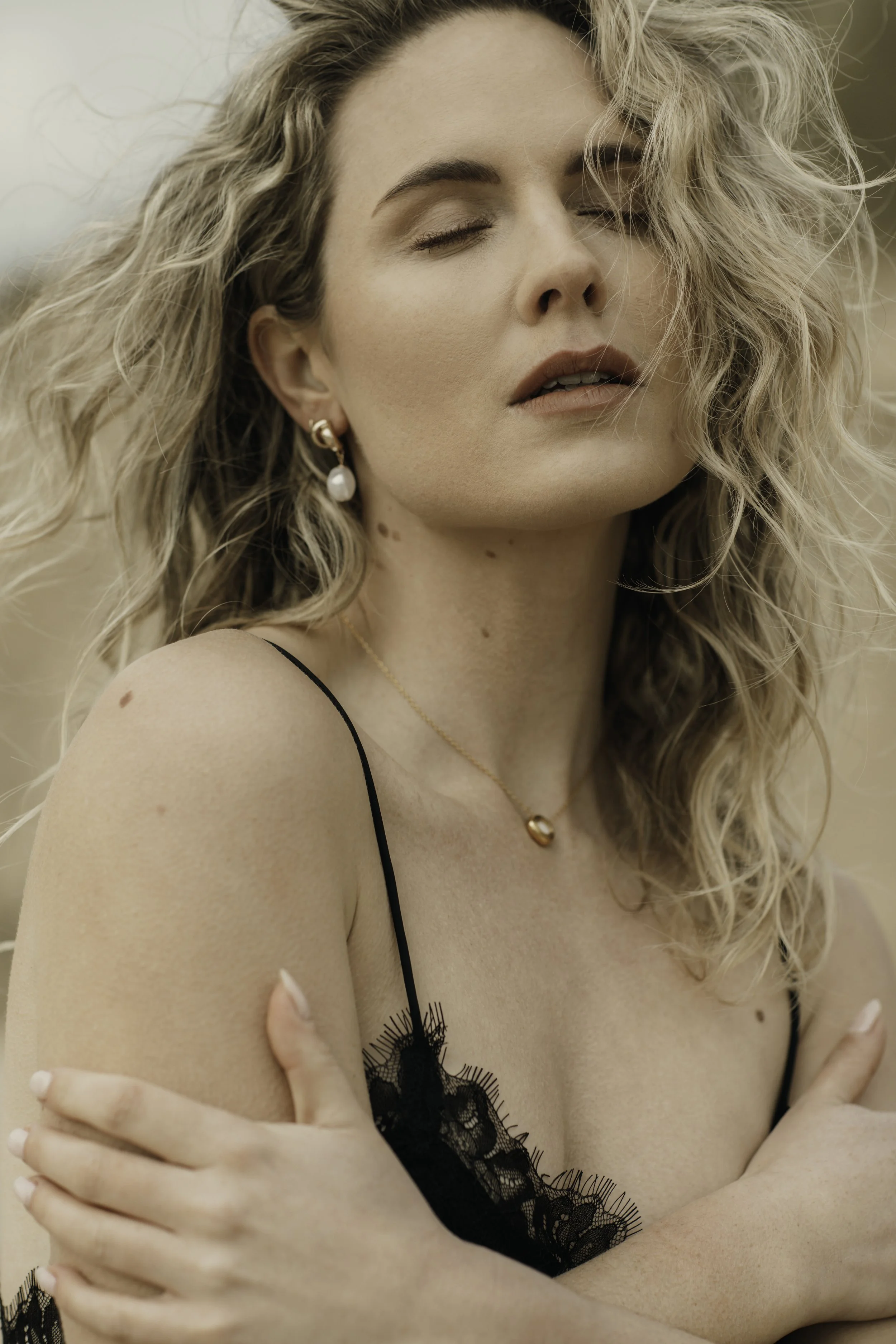 A woman with blonde curly hair is posing with her eyes closed and arms crossed over her chest. She is wearing a black lace lingerie top, pearl earrings, a gold necklace with a heart pendant, and is outdoors with a blurred natural background.