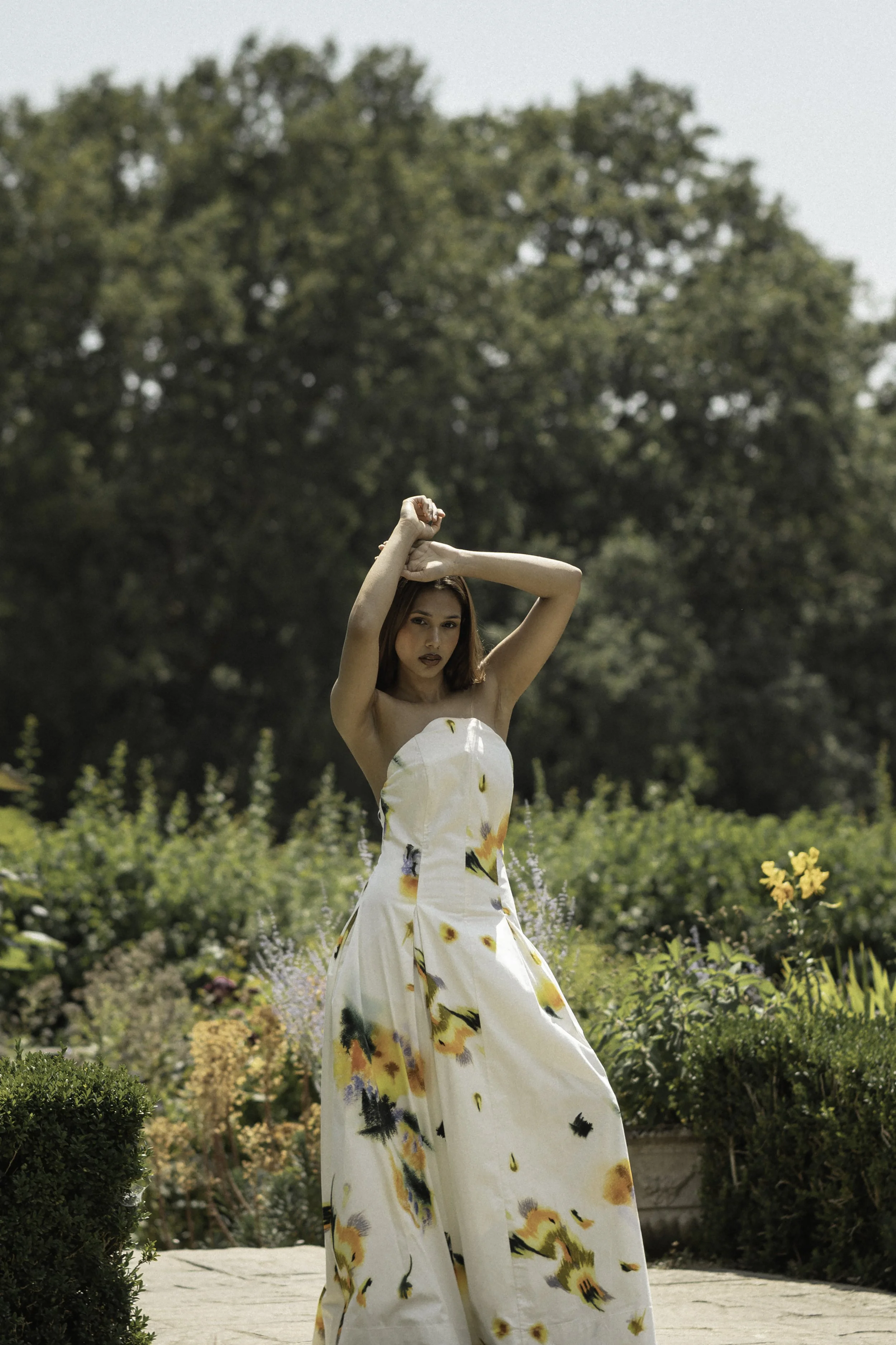 A woman in a strapless floral dress standing outdoors in a garden with lush greenery and flowers, holding her arms above her head.