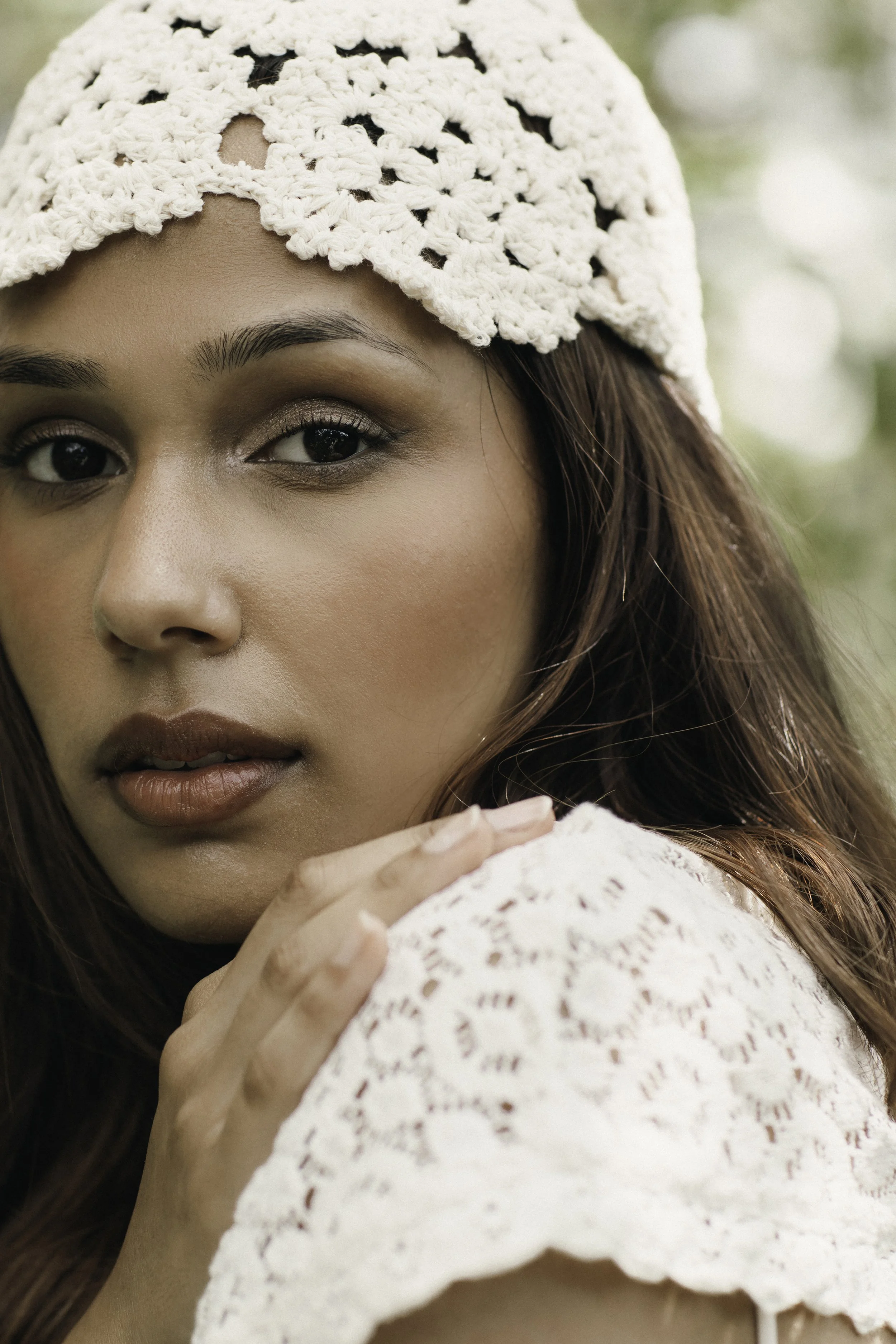 A close-up portrait of a young woman with long brown hair, wearing a white crochet hat and white lace top. She has makeup on with neutral tones and is holding her hand near her face with a blurred background of green foliage.