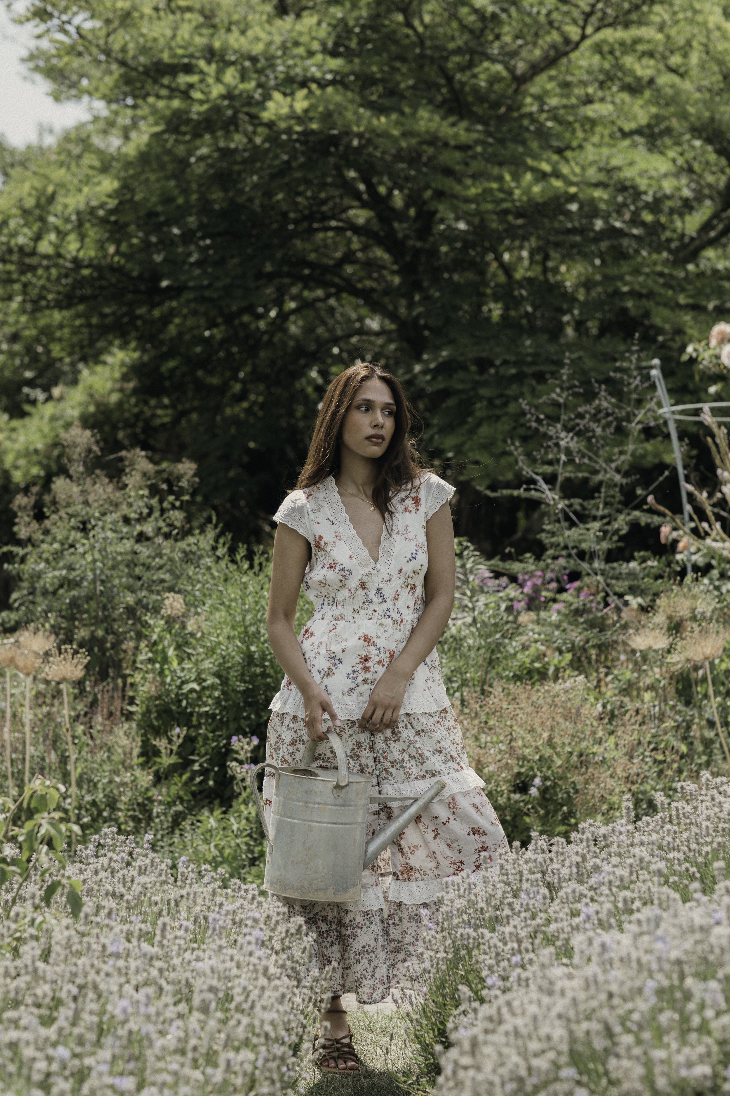 A woman in a floral dress holding a watering can standing in a garden filled with flowering plants and greenery.