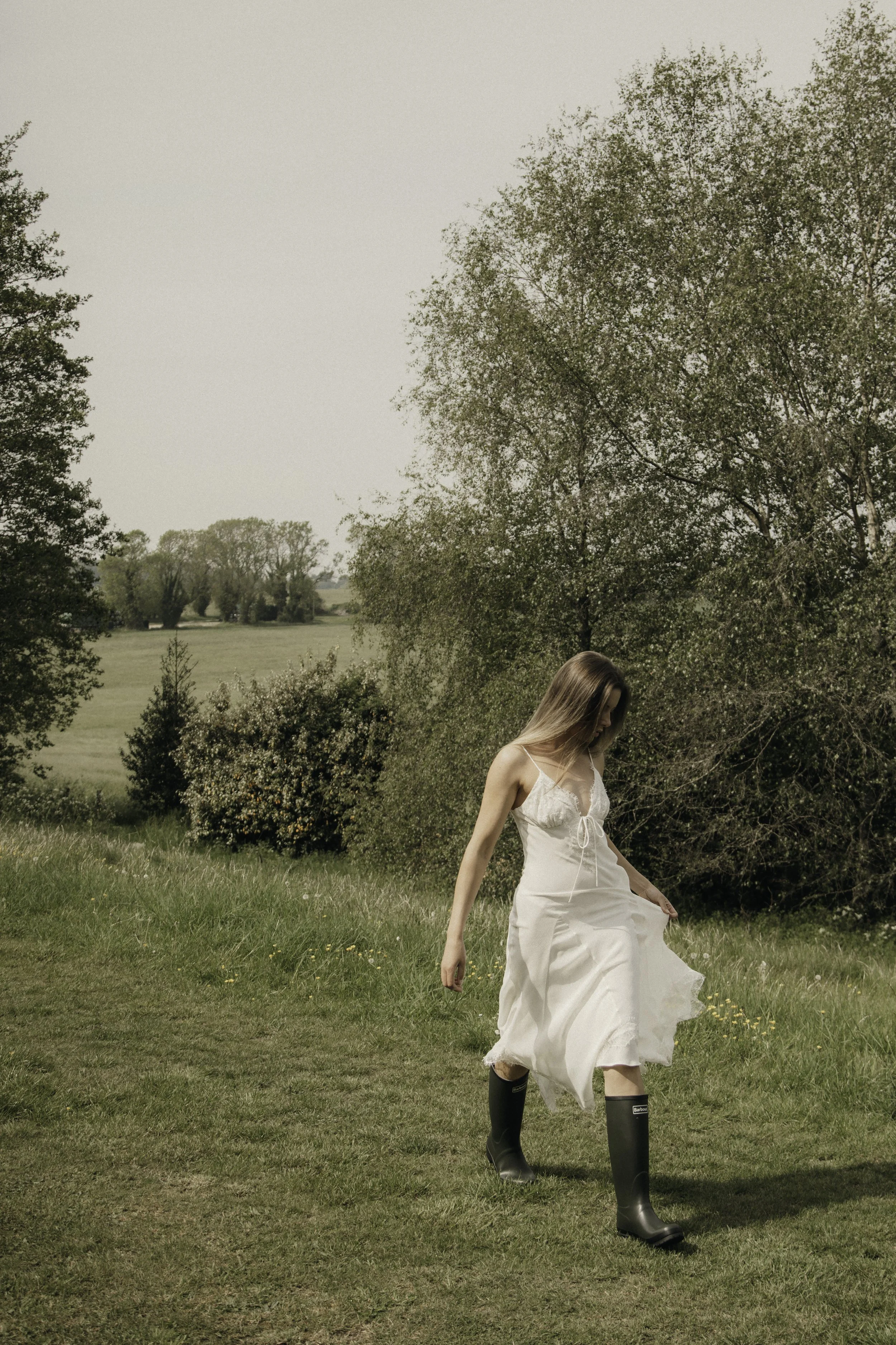 A woman in a white dress and black boots walking across a grassy field with trees and open land in the background during daytime.