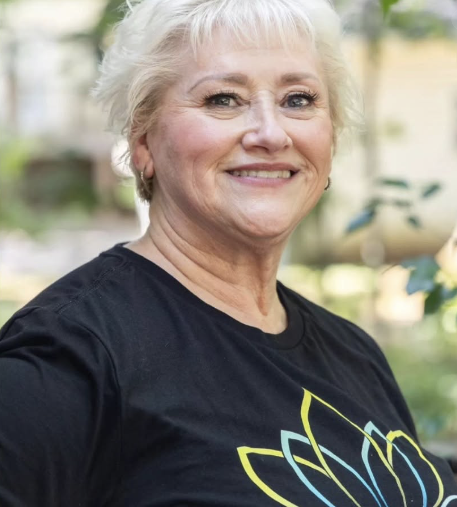 A smiling woman with short gray hair wearing a black T-shirt outdoors, with blurred green trees and plants in the background.