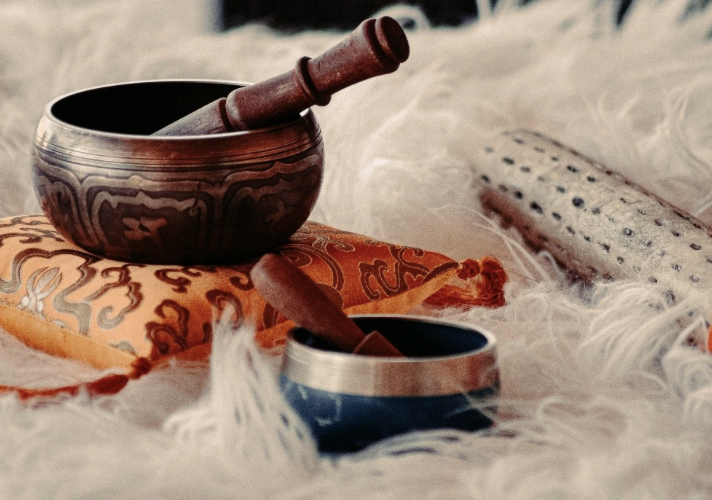 A Tibetan singing bowl with a wooden striker on a decorative fabric, with another smaller singing bowl nearby, and a white textured object in the background.