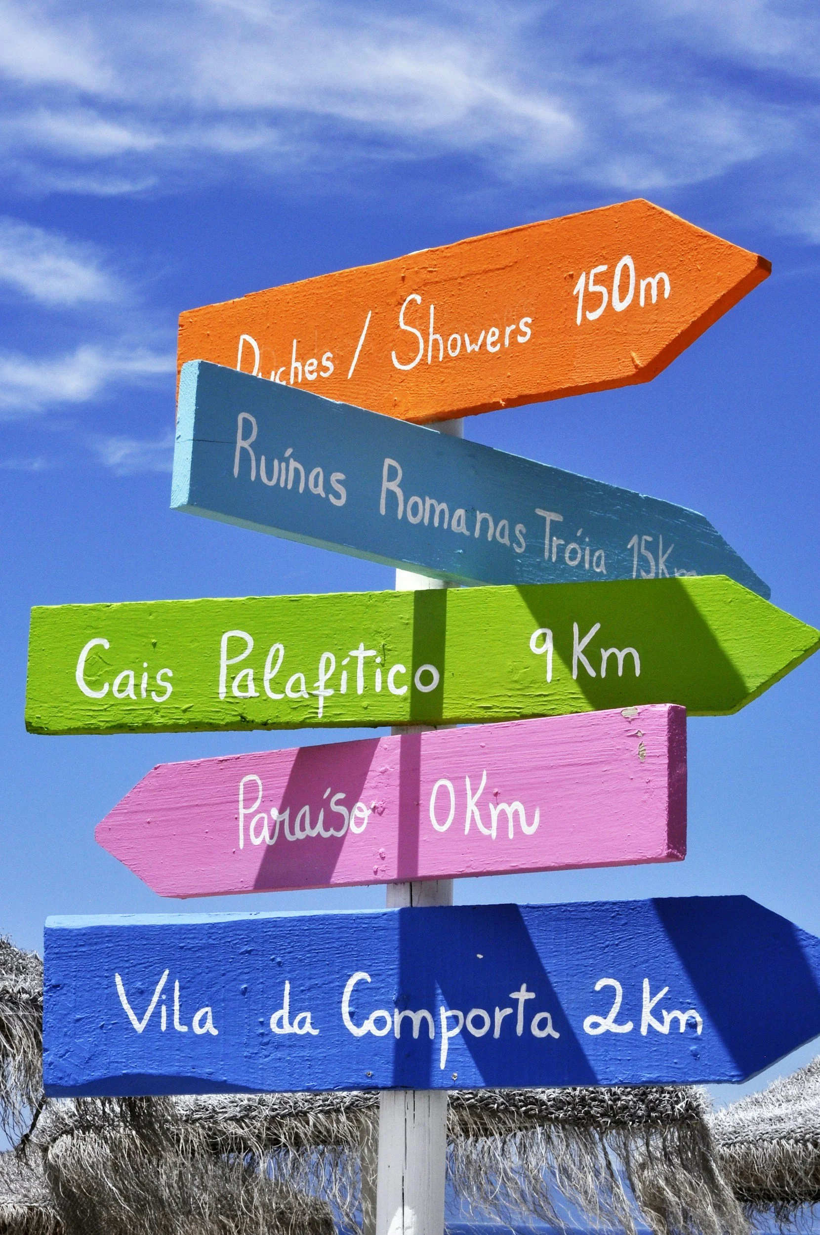 Colorful directional signs pointing to various locations with distances, against a blue sky with some clouds. The signs are painted in orange, blue, green, pink, and dark blue, with names and distances to places like Paraíso, Vila da Compostar, Ruínas Romanas Troia, Cais Palafítico, and others.