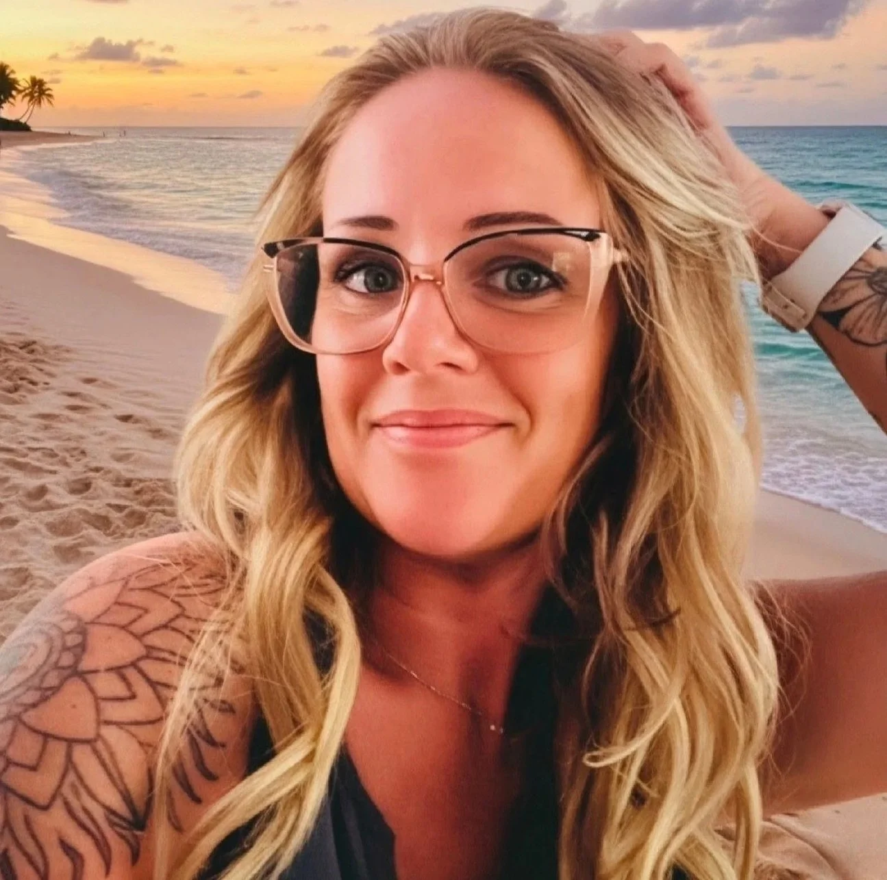 A woman with long blonde hair and glasses taking a selfie on the beach at sunset, with palm trees and ocean in the background.