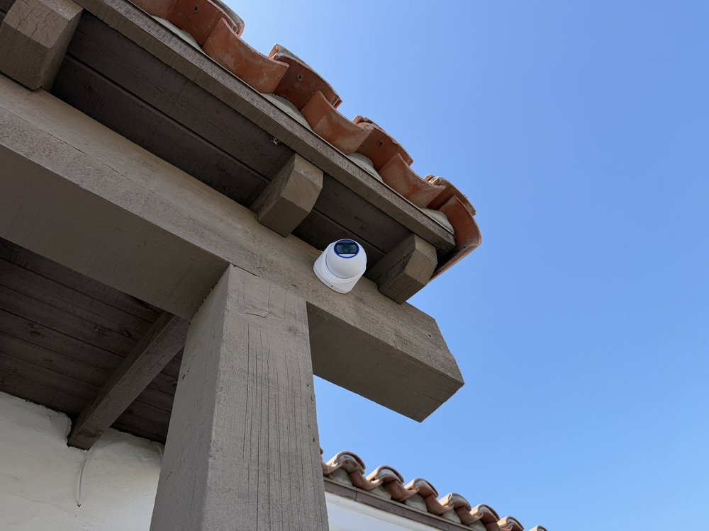White security camera mounted beneath a timber and tile roof against blue sky.