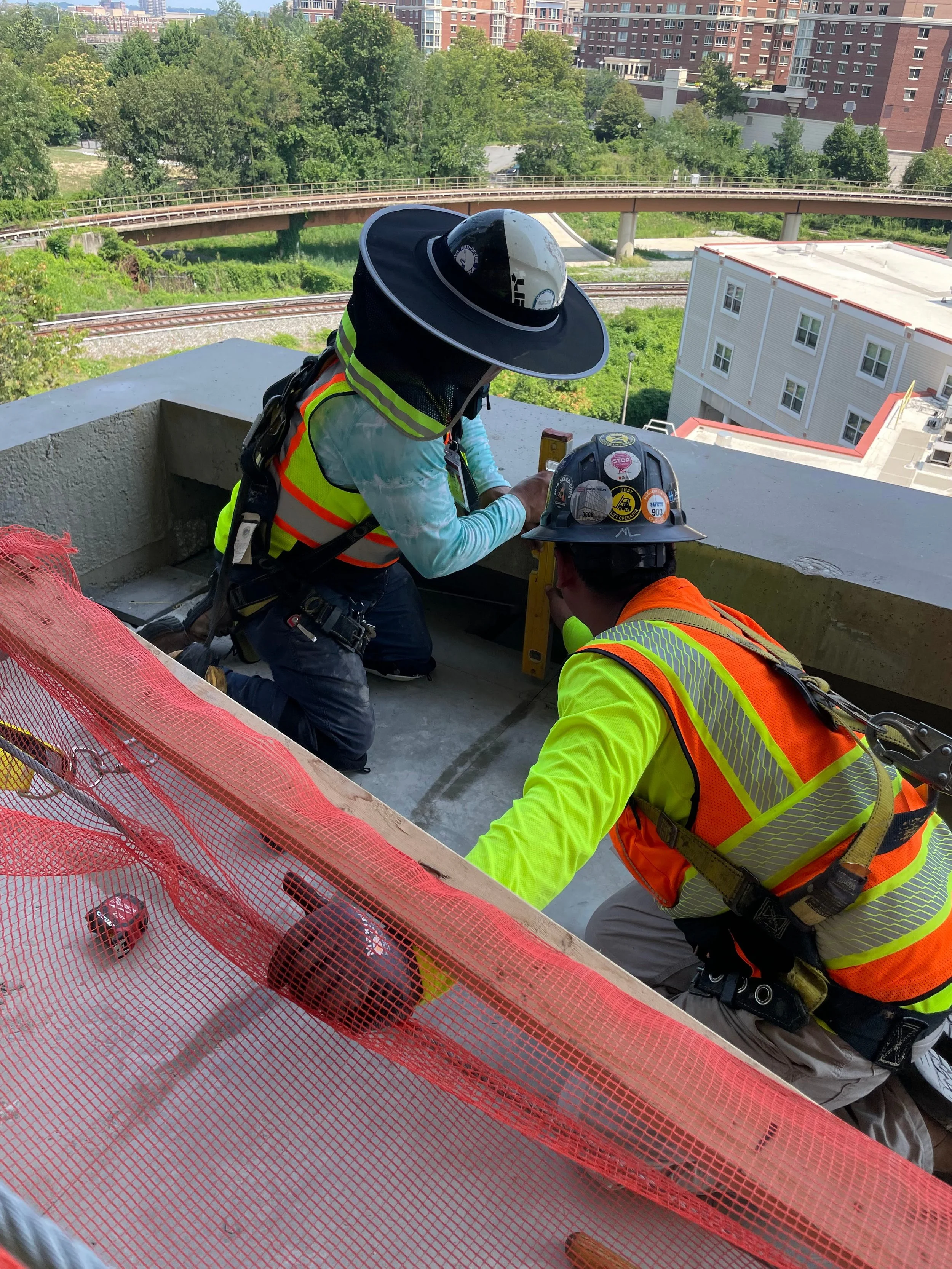 Two construction workers, wearing safety helmets and high-visibility vests, working on a building's rooftop.