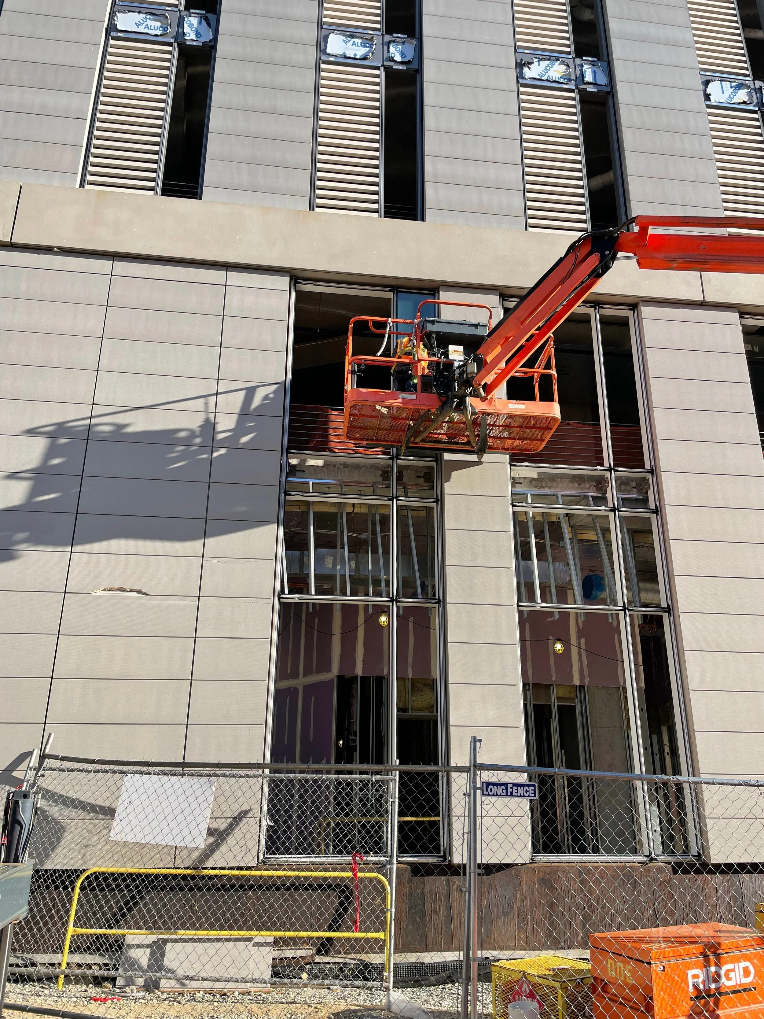 Construction site in front of a modern building with large glass windows and vertical louvers, featuring a cherry picker lift, a chain-link fence, and construction materials and equipment.