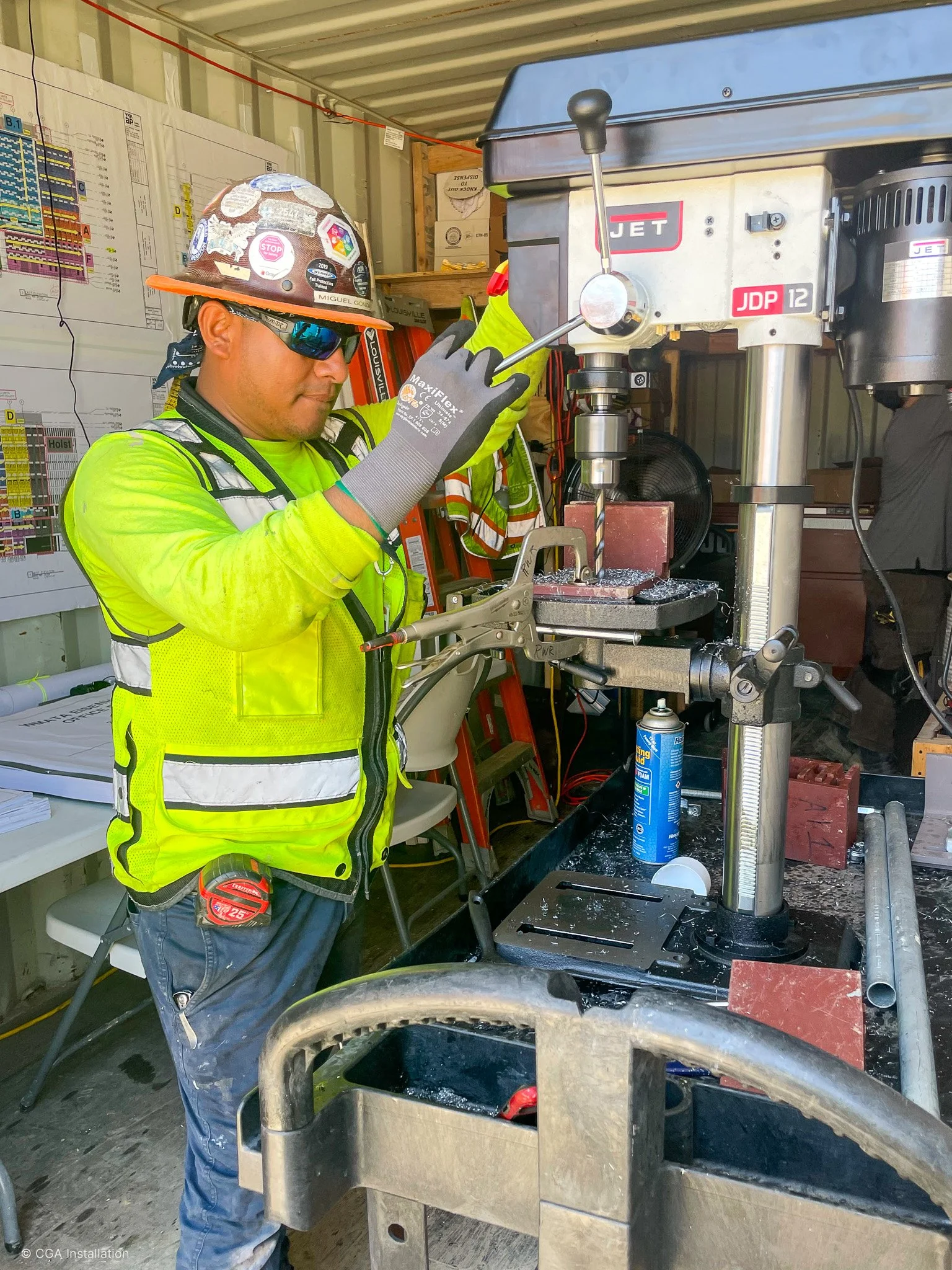 A construction worker in a yellow safety jacket, jeans, and a helmet with stickers, is working with a large drill press in a workshop. The worker is wearing safety gloves and glasses. The workshop has various tools, wiring, and safety posters on the 