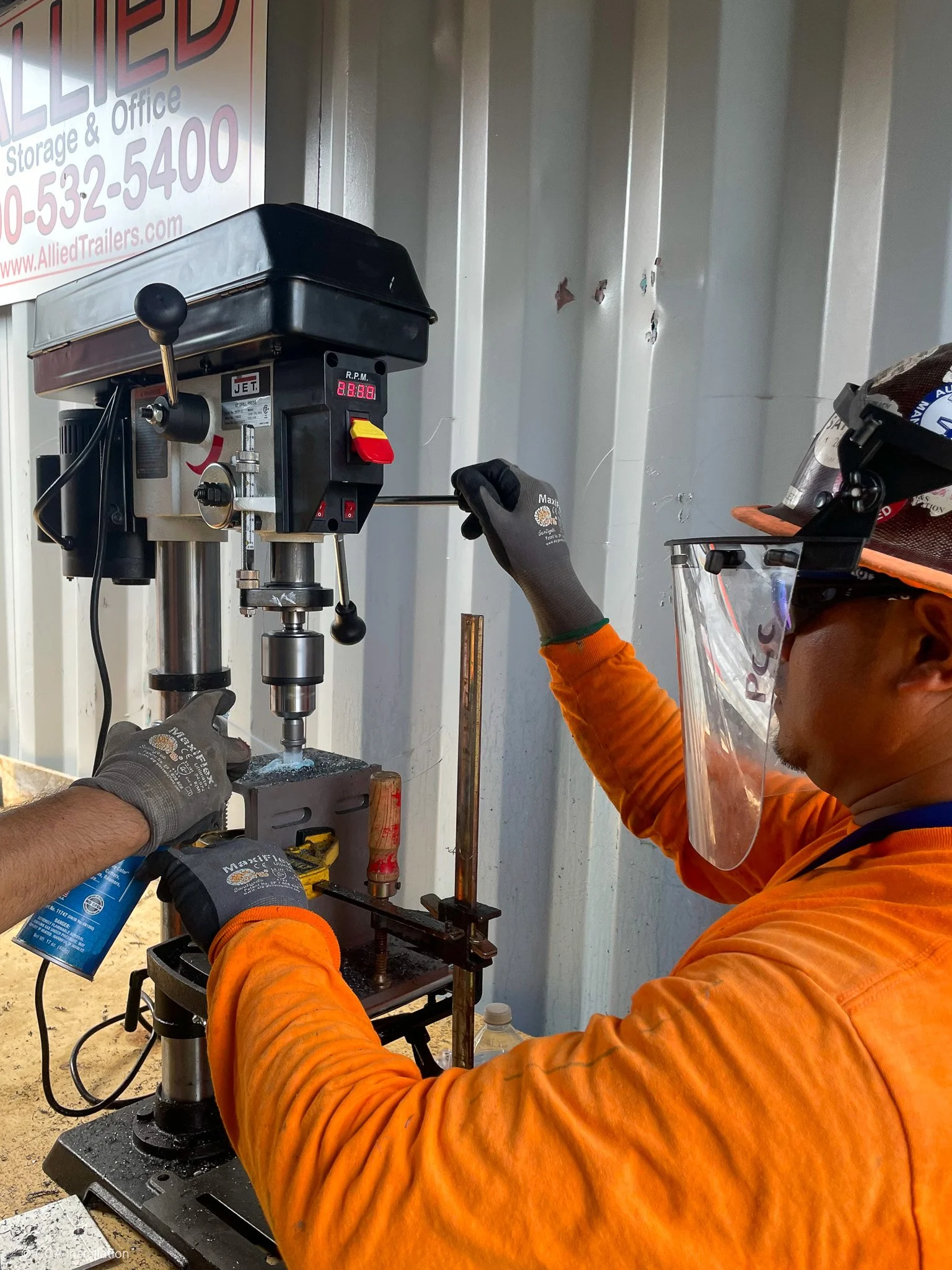 A person wearing gloves and protective gear is operating a drill press machine in an indoor workshop or storage area. The person is wearing an orange long-sleeve shirt and a helmet with a face shield. The drill press is positioned near a corrugated m