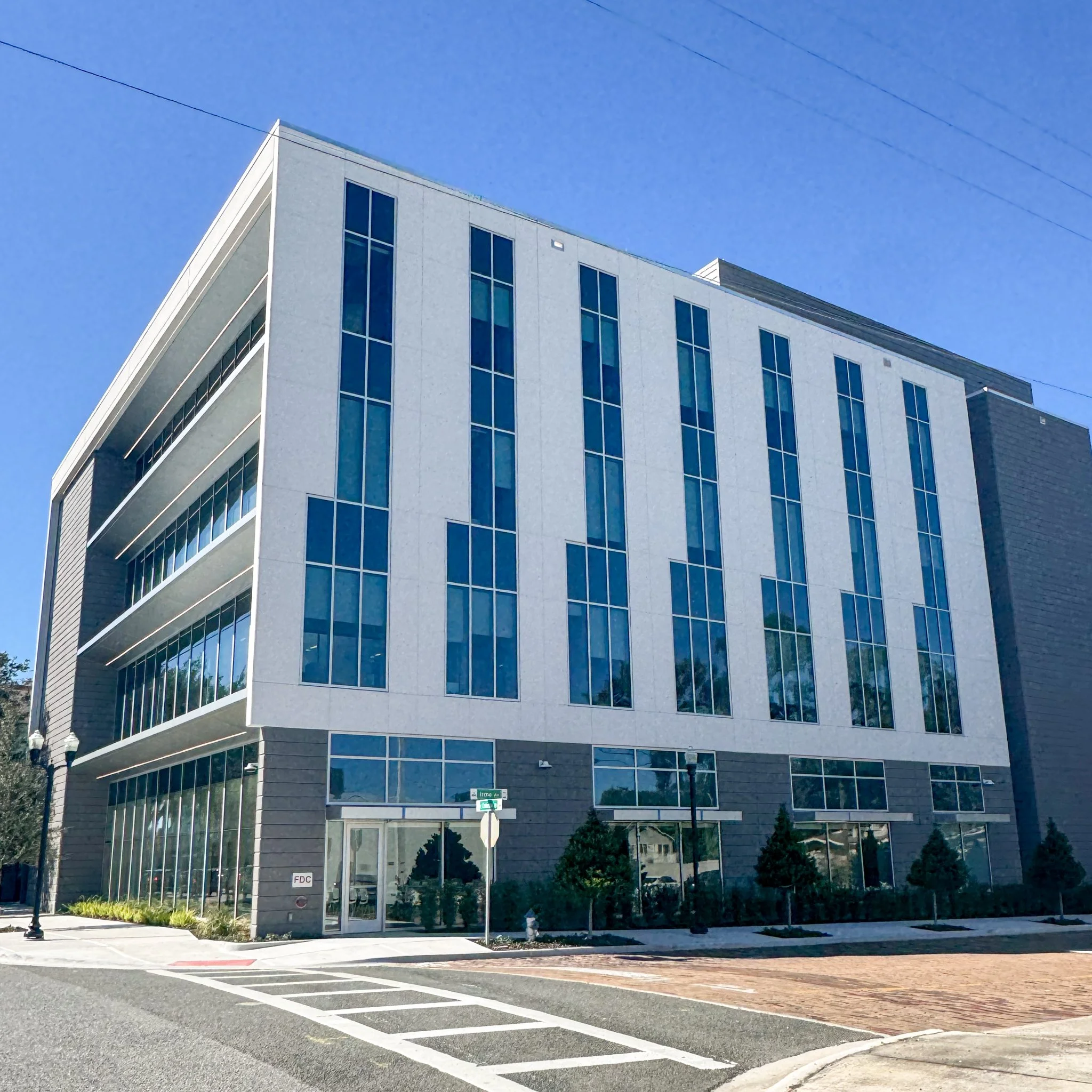 Modern multi-story building with large glass windows and a white exterior, situated on a city street with crosswalks and street signs, under a clear blue sky.