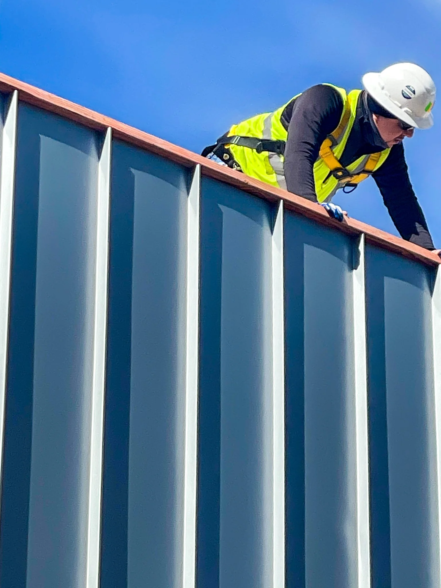 A construction worker wearing a safety helmet and a neon yellow vest is working on the roof of a building with vertical metal siding, under a clear blue sky.