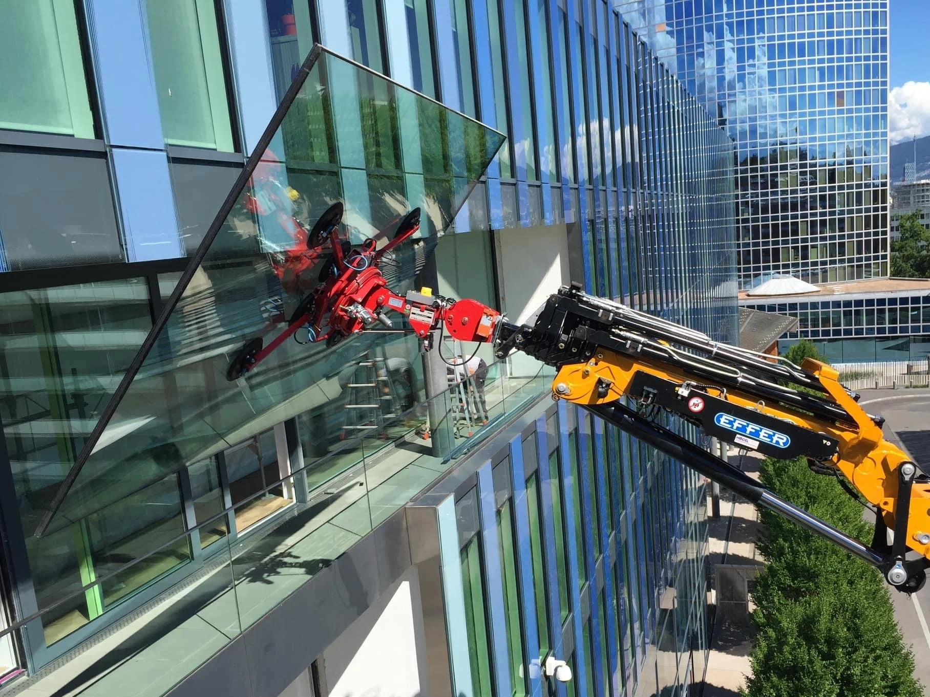 A telescopic crane is installing a large glass window on a modern multi-story glass building with reflections of the sky and clouds. The crane's arm extends to reach the window, and a worker is visible inside the building.