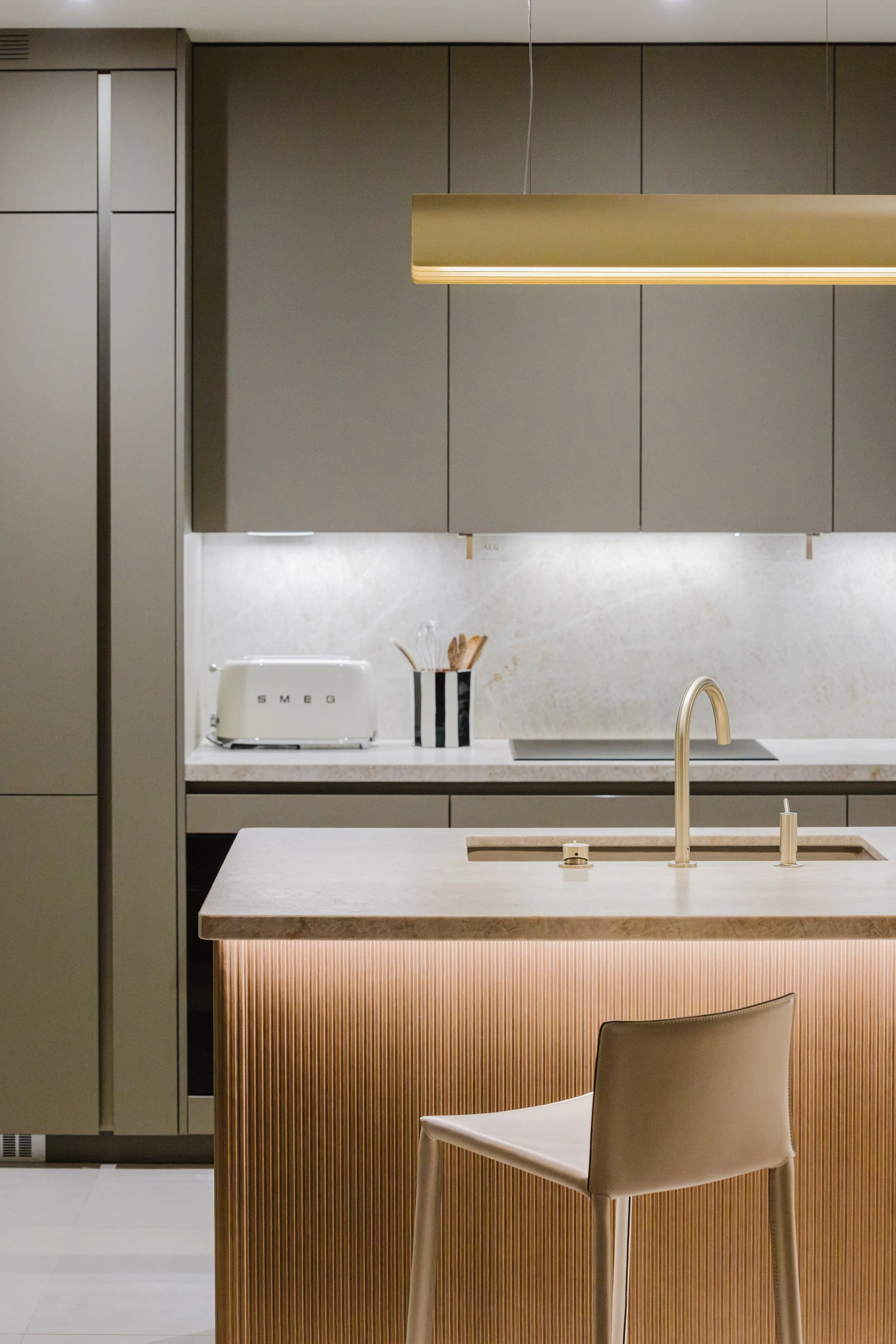 Modern kitchen with beige cabinetry, a marble countertop, a gold-colored pendant light, a white SMEG toaster, a striped black and white utensil holder, a beige chair, and a gold sink faucet.