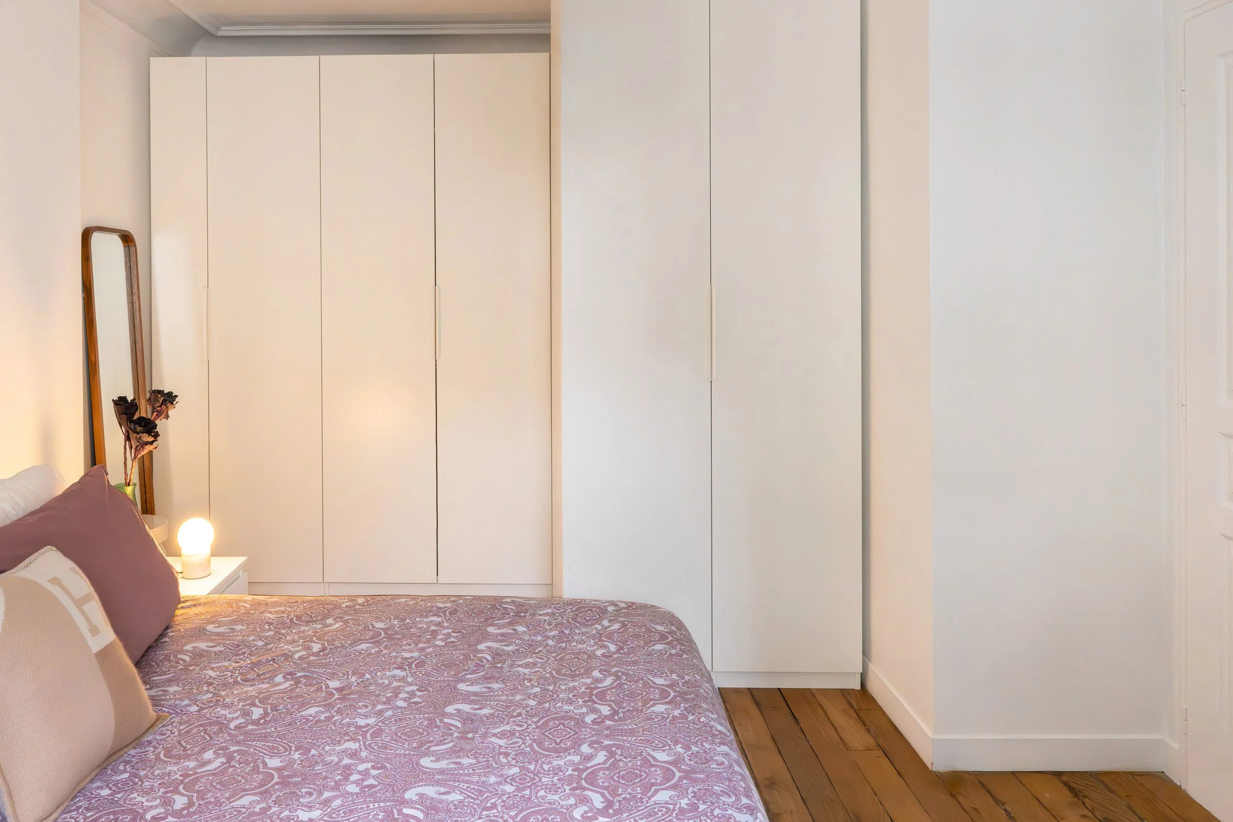 A bedroom featuring a bed with pillows, a patterned bedspread, a bedside table with a lit lamp, and a large white wardrobe. There is a mirror and a vase with flowers on the bedside table.