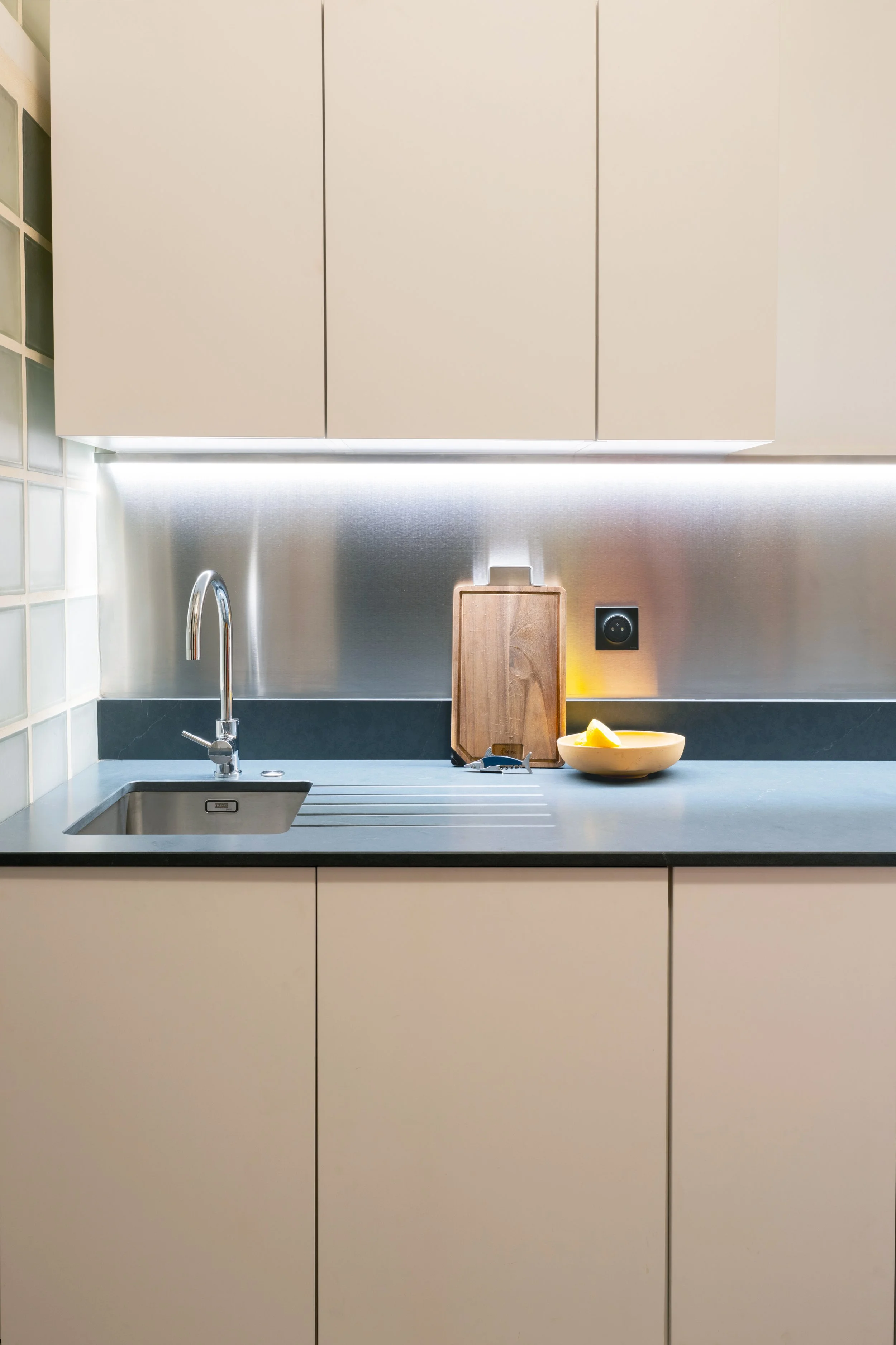 Modern kitchen with white cabinets, a stainless steel backsplash, a sink with a faucet, a wooden cutting board, a bowl with lemons, and a power outlet.