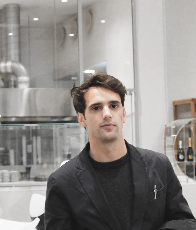 A young man sitting in a modern cafe, wearing a black blazer over a black shirt, with a neutral expression, in front of a counter with champagne bottles.