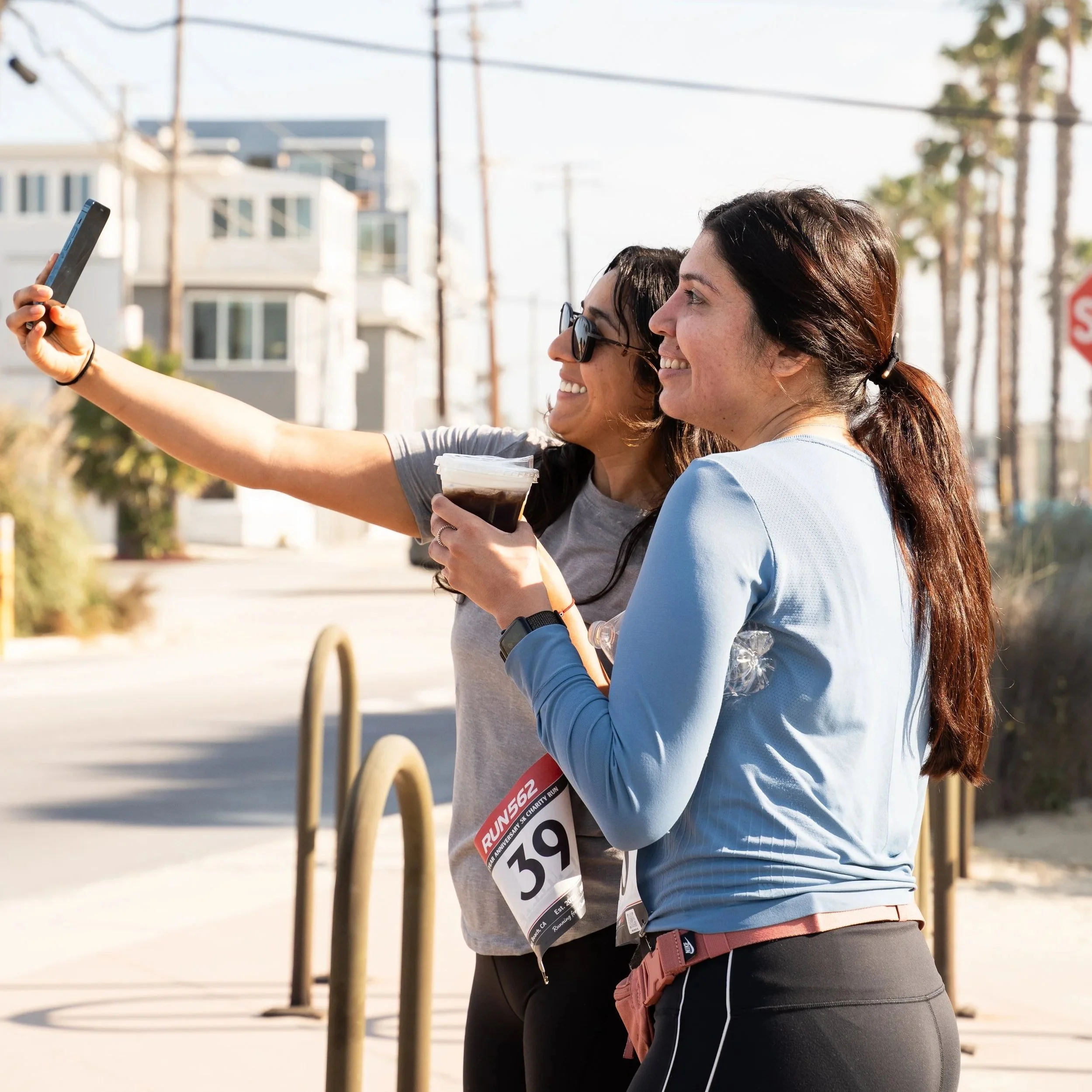 Two women participating in a running event, taking a selfie together on a sunny day, wearing race bibs and athletic clothing.