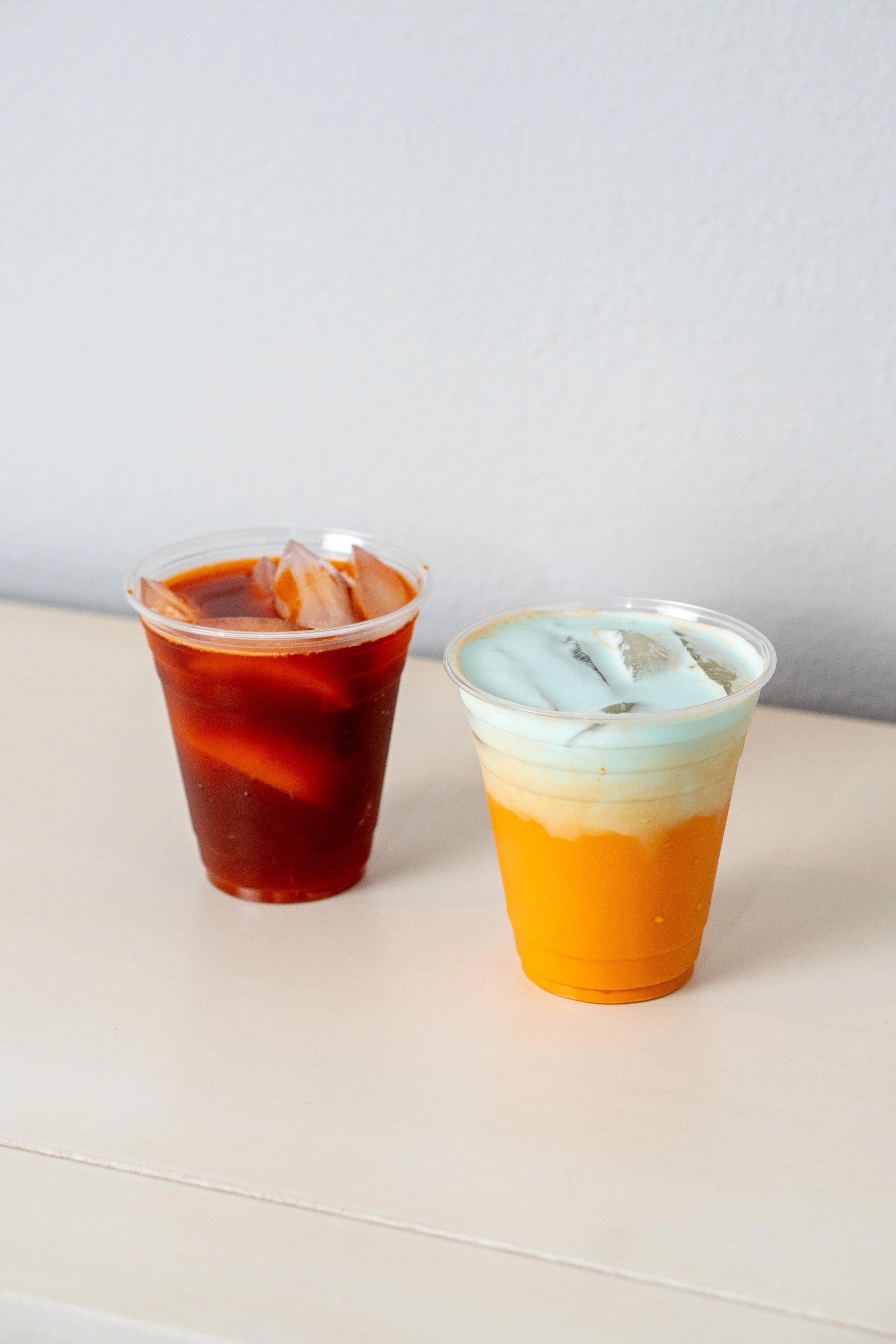 Two cups of beverages on a white surface, one with a red drink and ice, and the other with a layered yellow, white, and blue drink with ice.