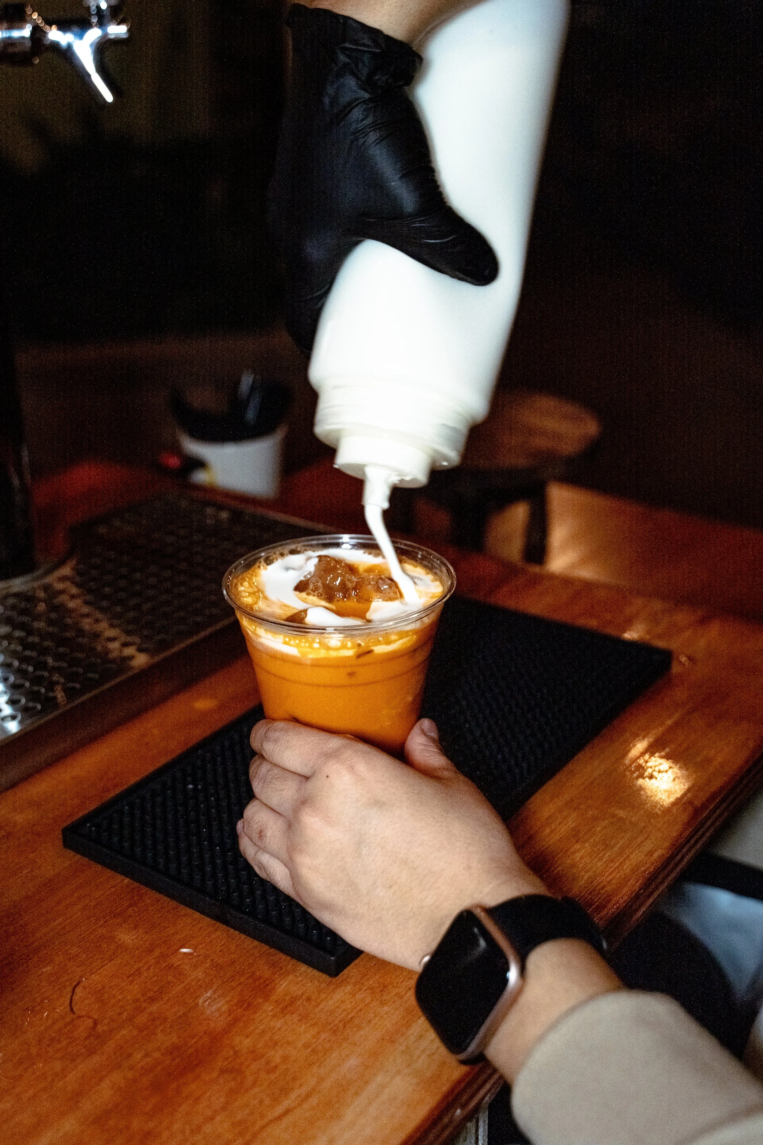 A person wearing a black wristwatch and a beige long-sleeve shirt is holding a clear plastic cup filled with iced coffee, with cream being poured into it from a white squeeze bottle. The person is using their other hand to hold the cup. In the background, there is a dark wood table with a black rubber mat on it, and some blurred objects on the table.