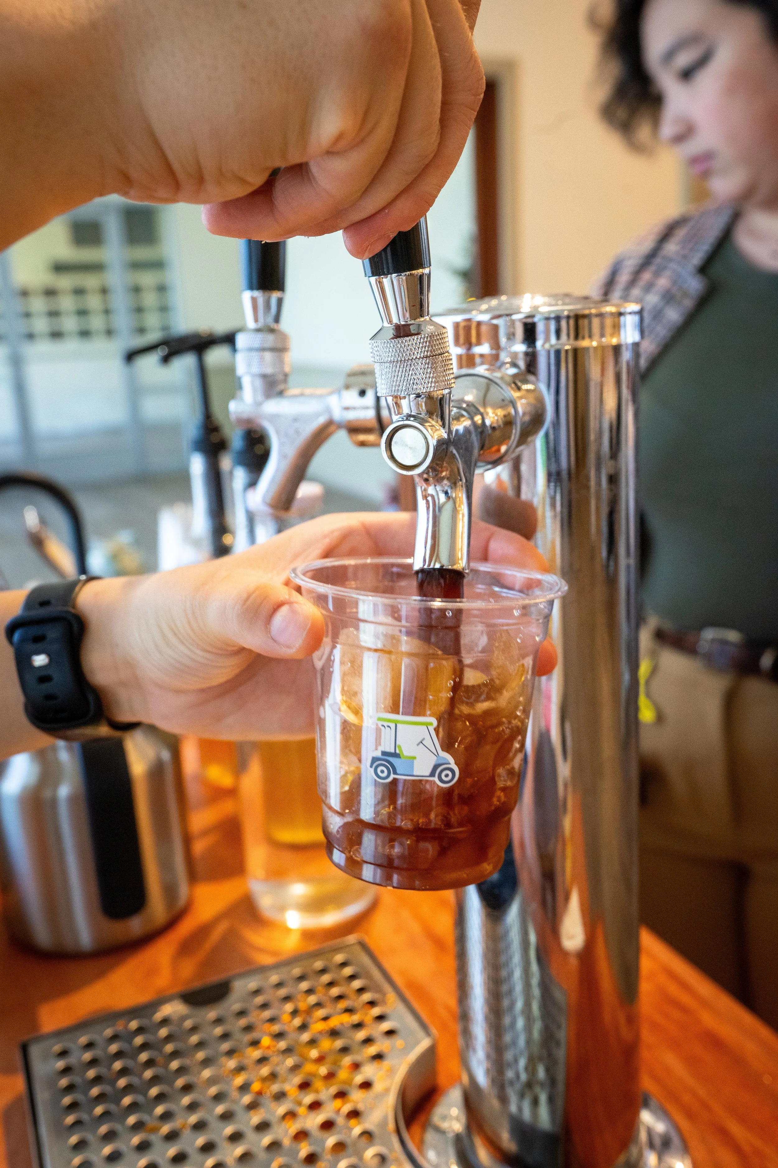 Person filling a plastic cup with iced tea from a tap at a self-serve station.