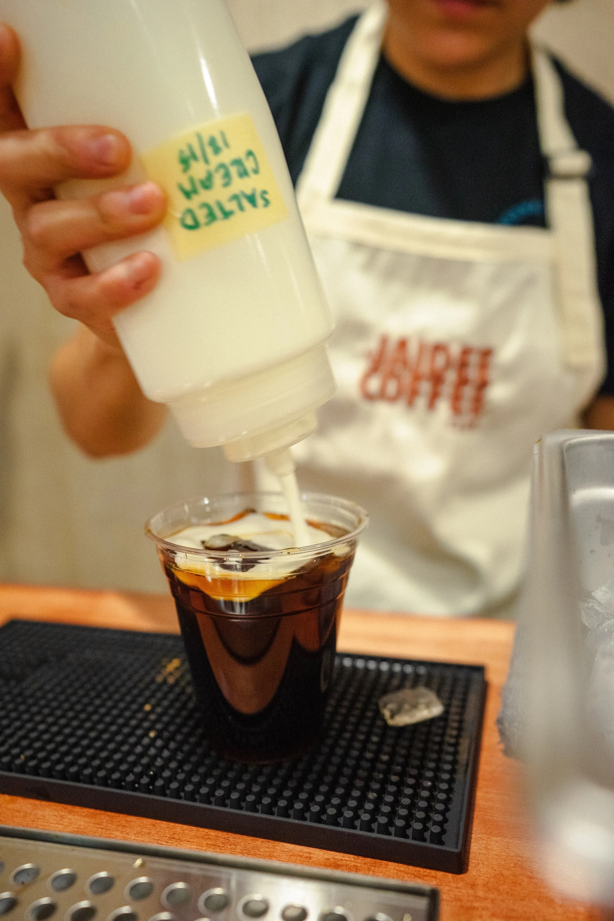 A person is pouring cream into a glass of iced coffee on a black mat on a wooden table.