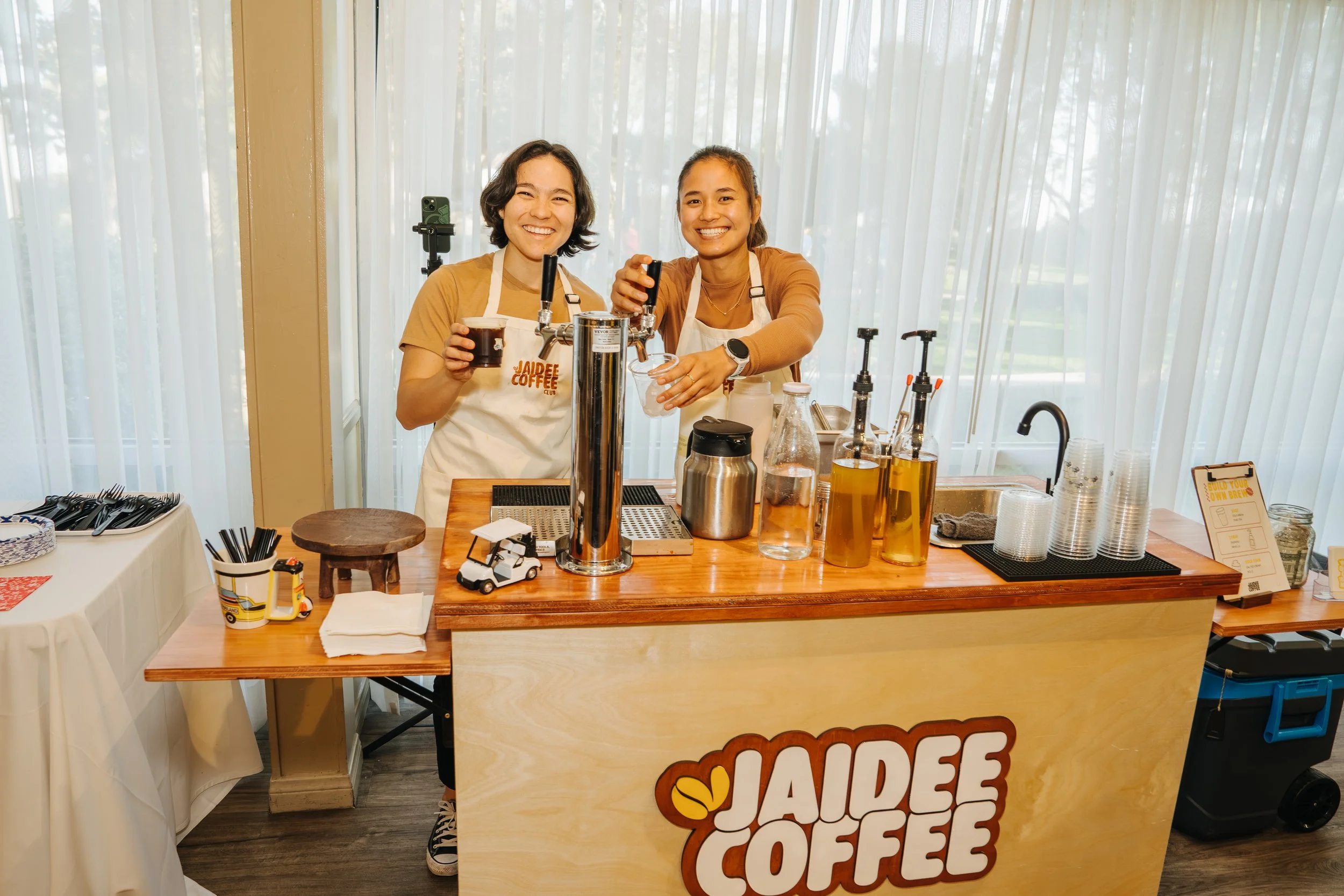 Two women smiling behind a coffee stand with various bottles and equipment, during daytime.