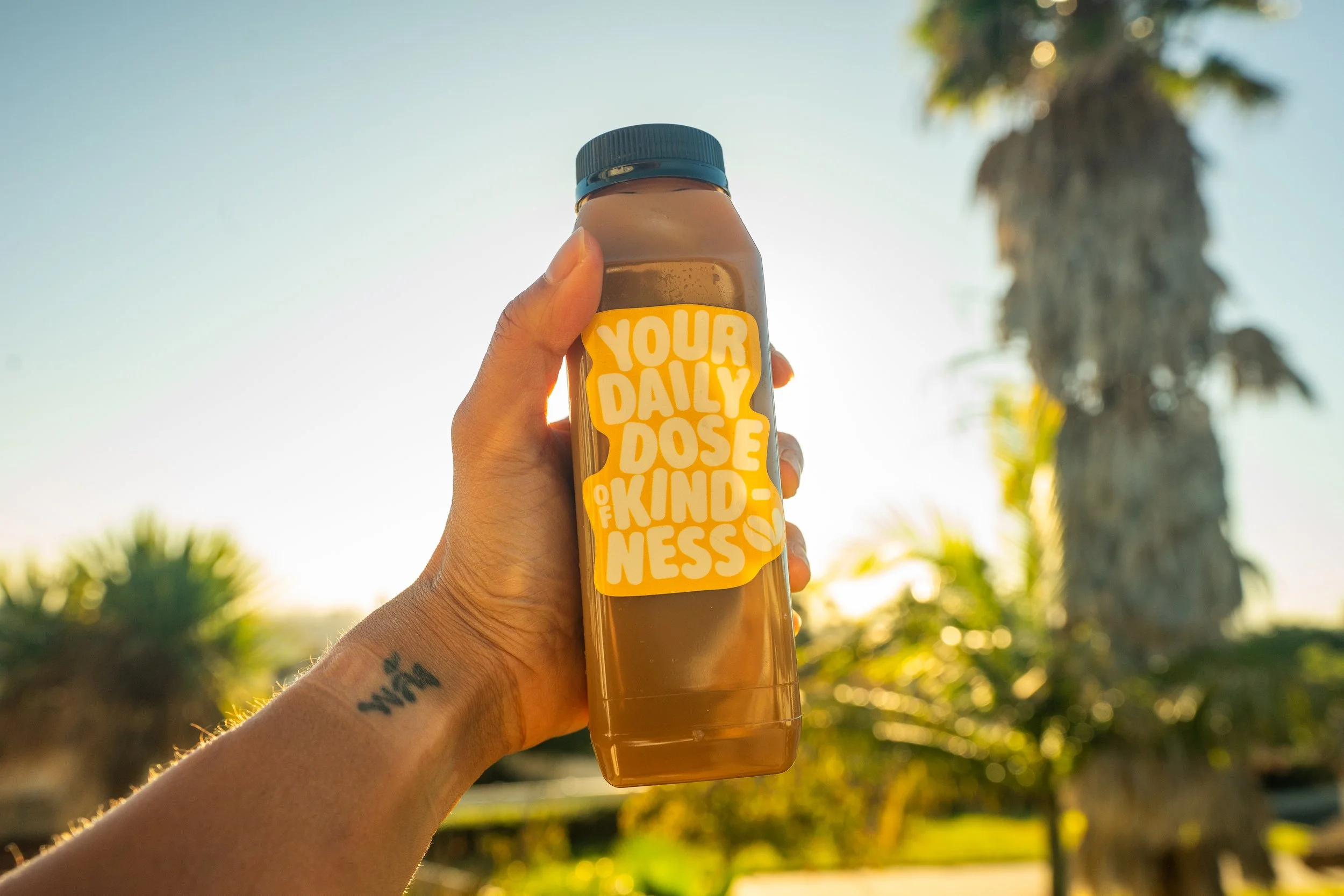 A person holding a bottle with a yellow label that reads 'Your Daily Dose of Kindness' against a sunny outdoor background with trees.