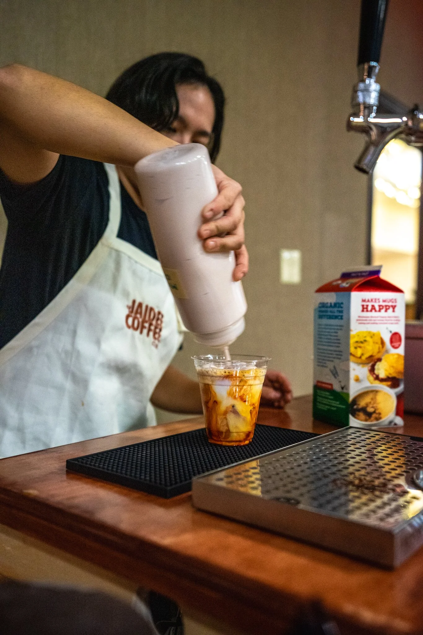 A person in a hamburger restaurant apron pouring a white liquid into a clear plastic cup with ice and caramel syrup, with a milk carton and an ice machine on a wooden counter.
