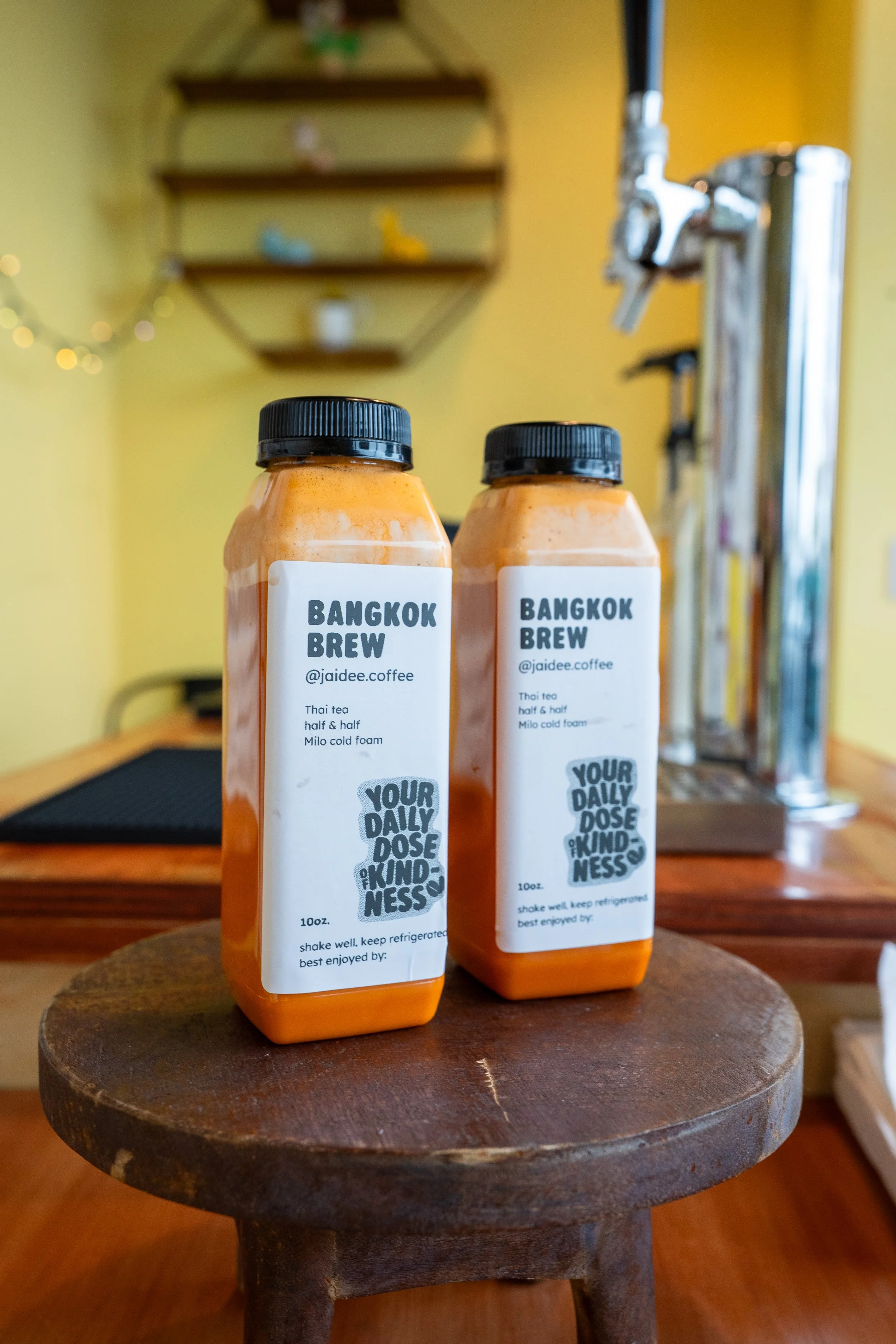 Two bottles of Bangkok Brew Thai tea with half & half and Milo cold foam on a wooden stool, in a cozy indoor setting with blurred background.
