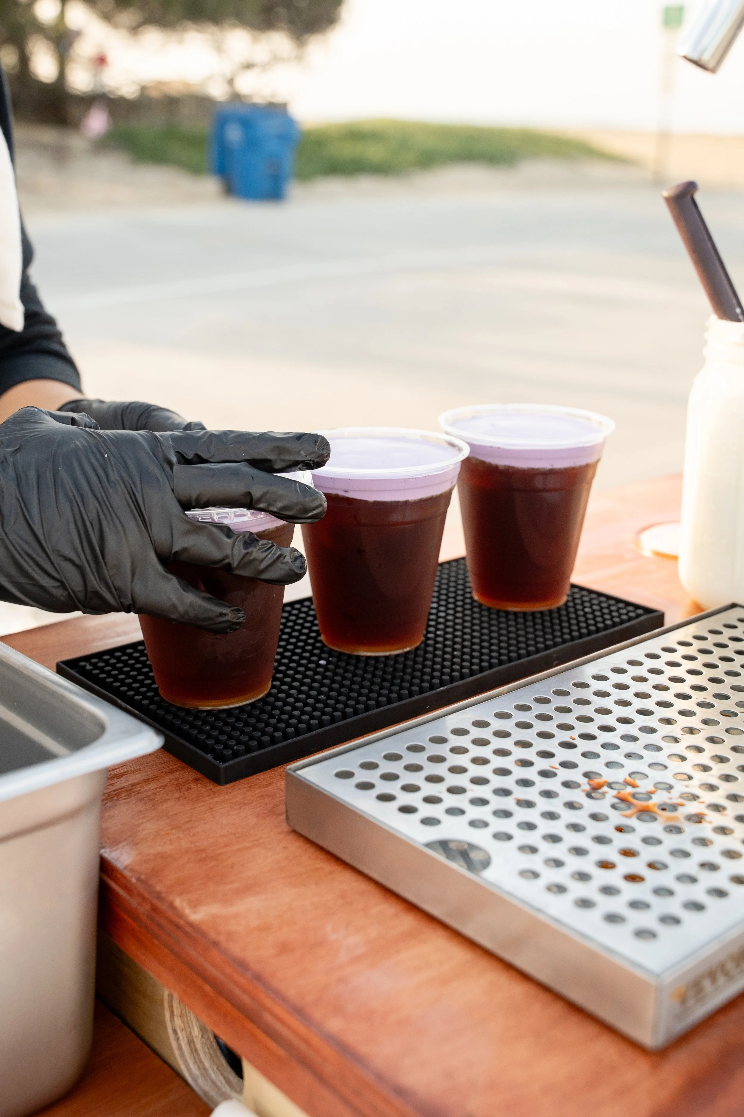 Person wearing black gloves preparing small cups of dark iced beverage outdoors, with a metal tray and a container of liquid nearby.